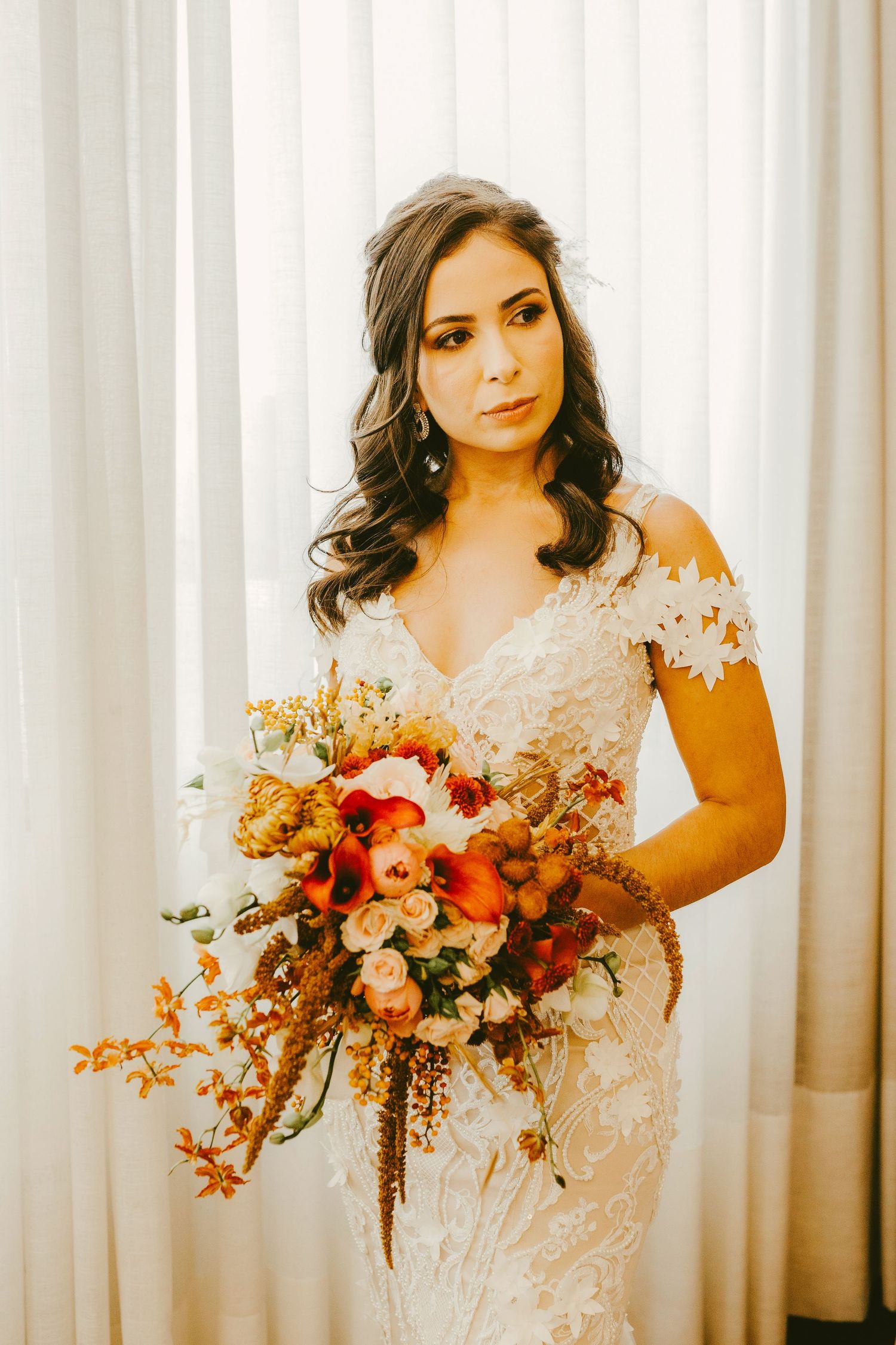 Bride in lace off-shoulder gown holding autumn bouquet with red and orange blooms, standing by white curtains.