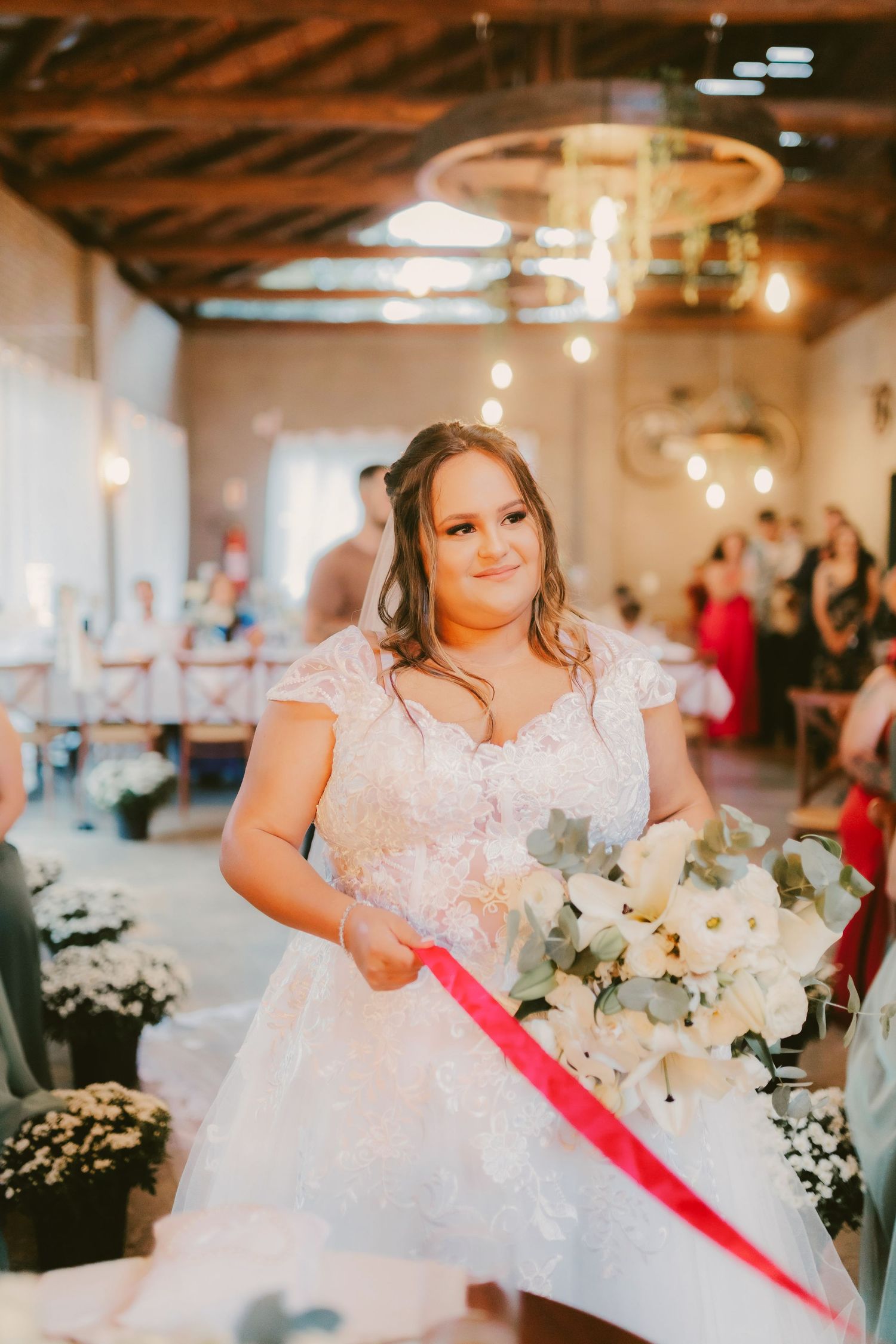 Bride in lace wedding dress holding white floral bouquet walks down aisle in rustic venue with warm string lights.