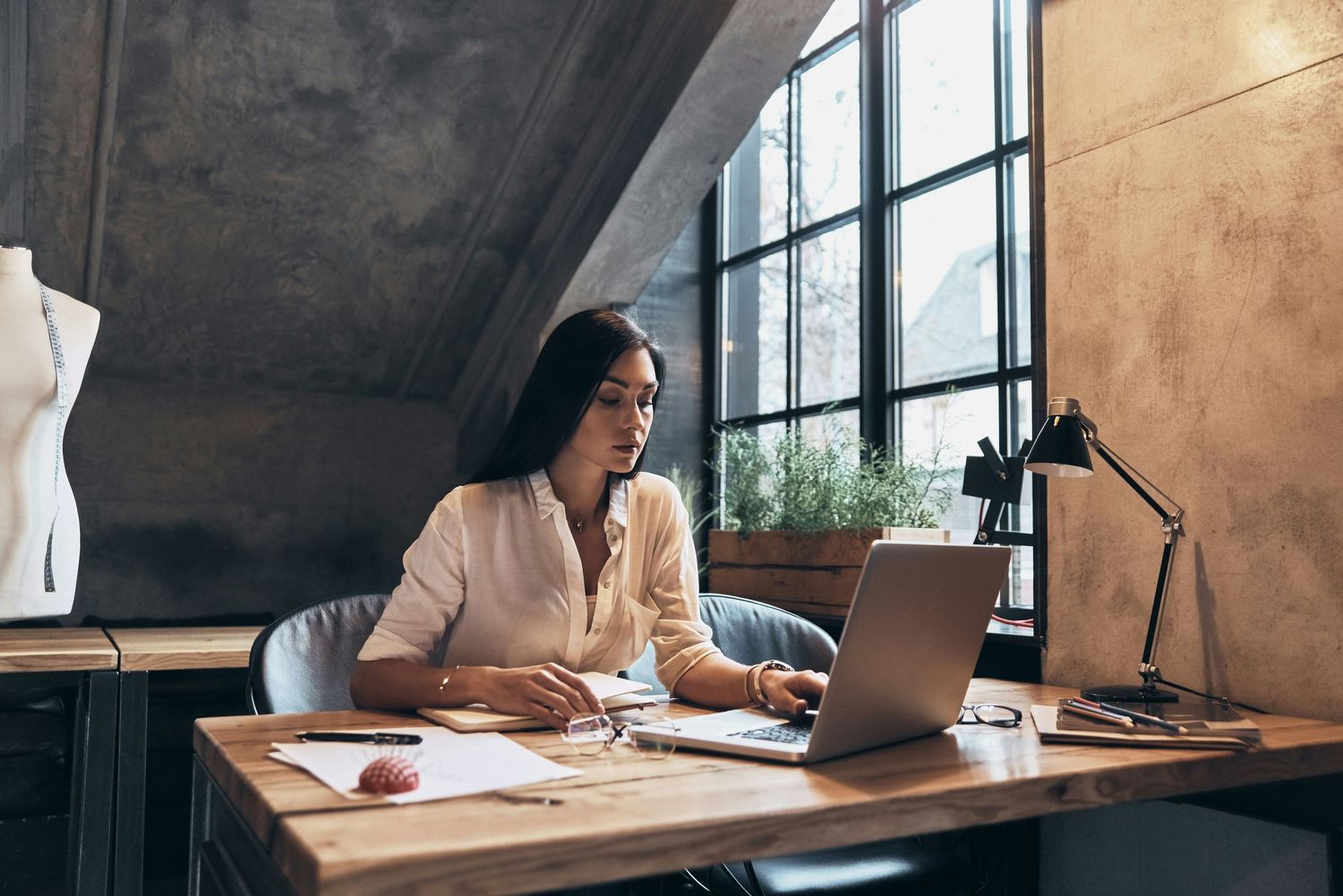 Professional woman in glasses working on laptop at wooden desk in industrial loft workspace.
