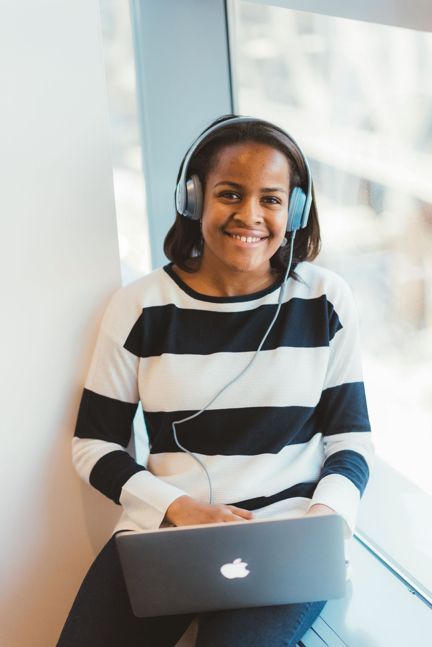Happy Black woman wearing striped shirt and headphones using Apple MacBook laptop by window.