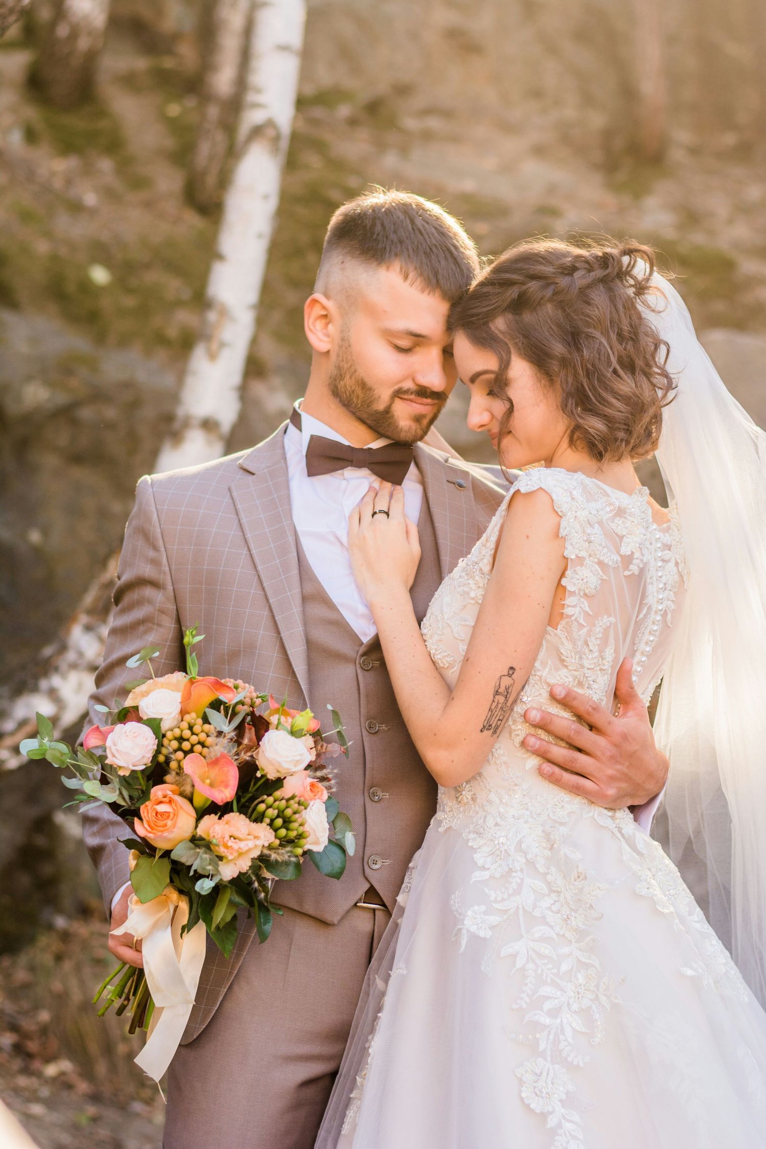 Bride and groom in beige suit embracing tenderly outdoors, holding colorful floral bouquet in sunlight.
