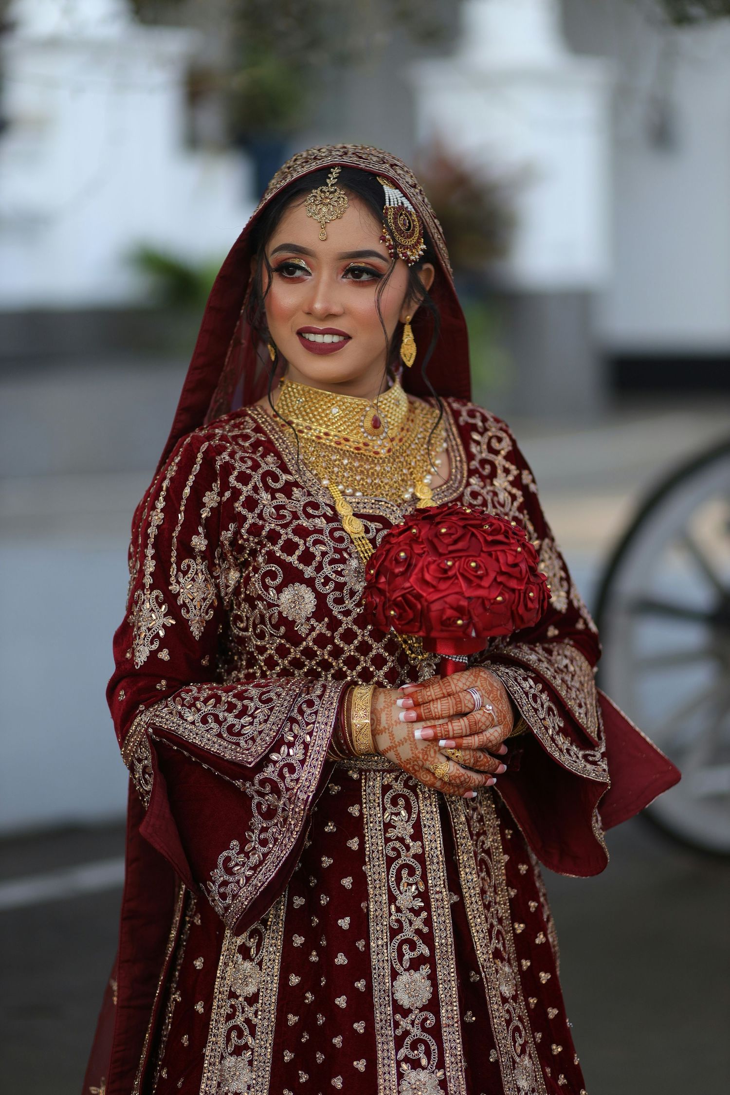 Indian bride in red and gold bridal lehenga holding red rose bouquet with traditional jewelry and mehndi.