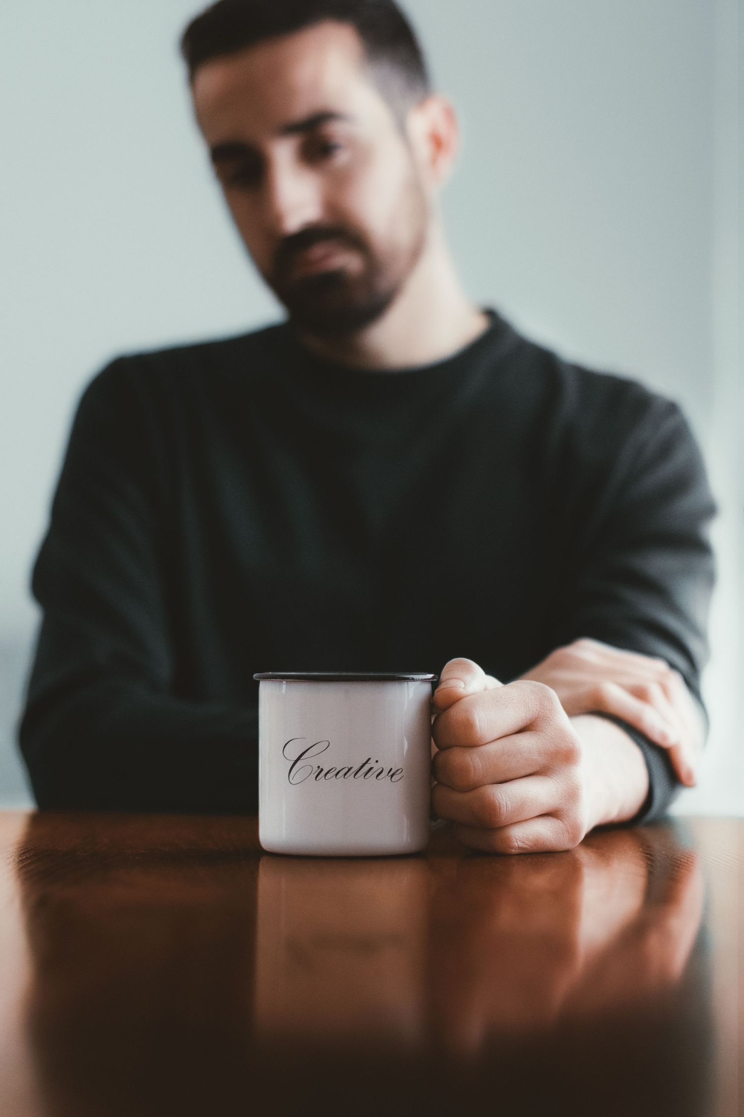 Bearded man in black sweater holding a mug labeled Creative at wooden table.