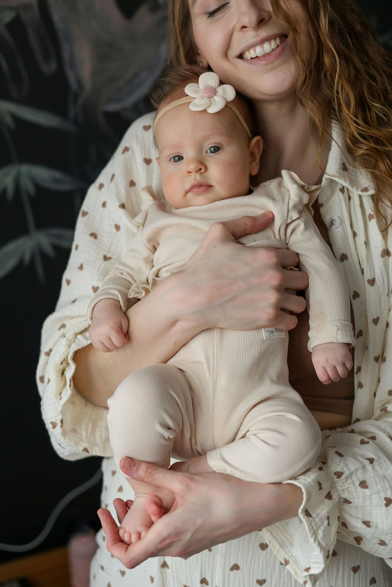 Smiling mother in cream heart-print dress holding newborn baby with flower headband.