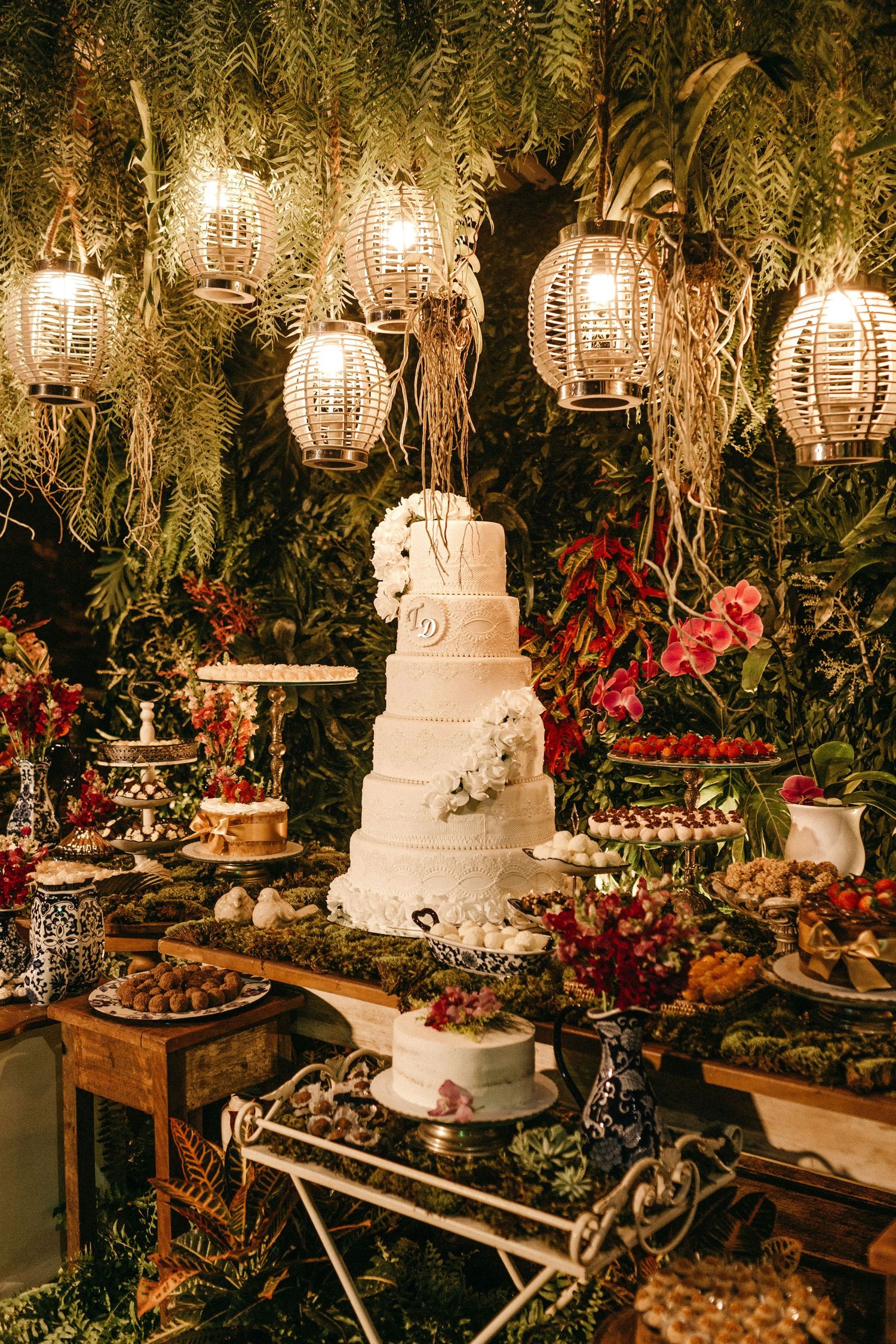 Lavish wedding dessert table featuring a tall white tiered cake surrounded by flowers and hanging lanterns.