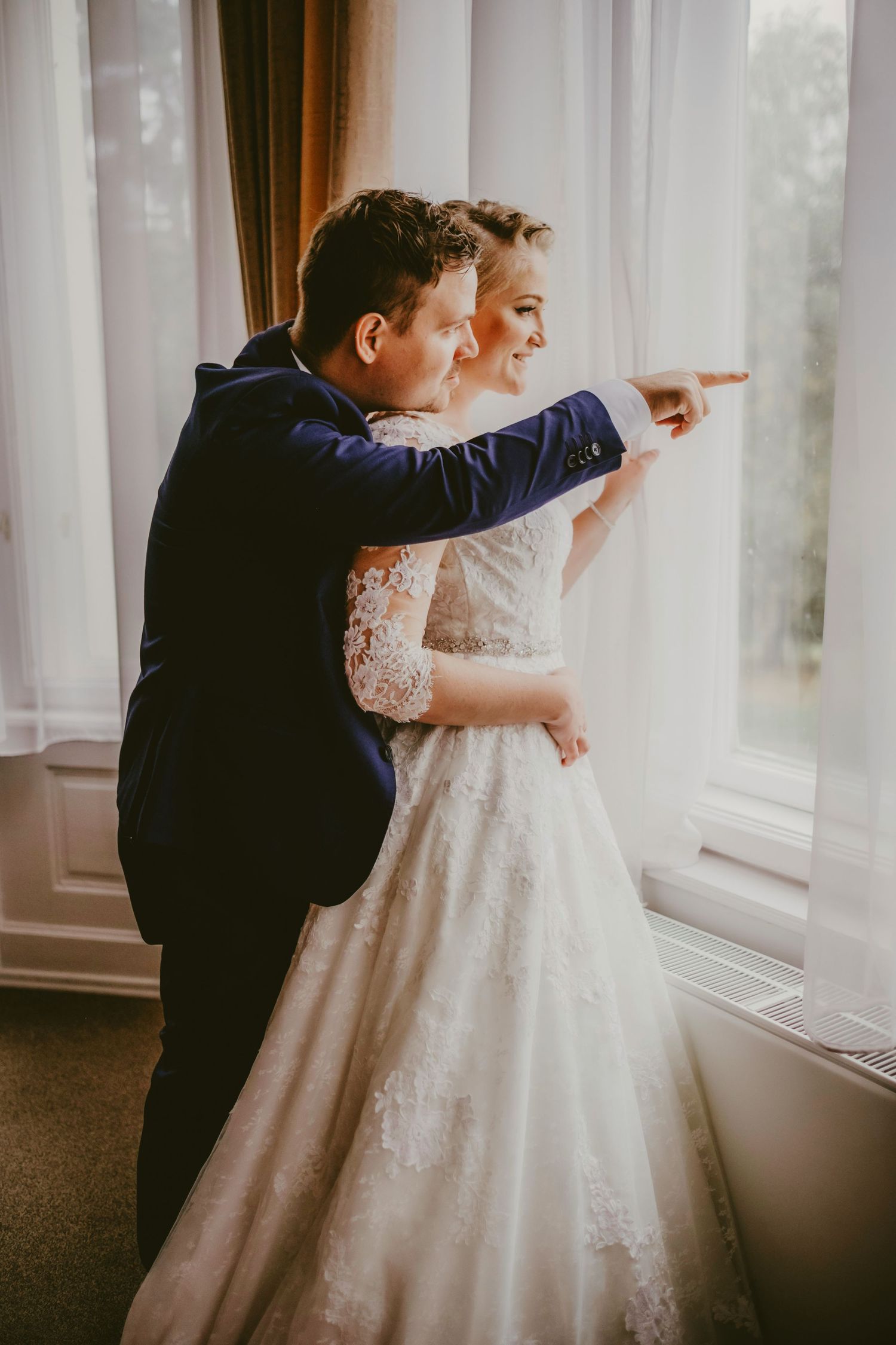 A bride in lace gown and groom in navy suit stand by window, pointing outside together on their wedding day.