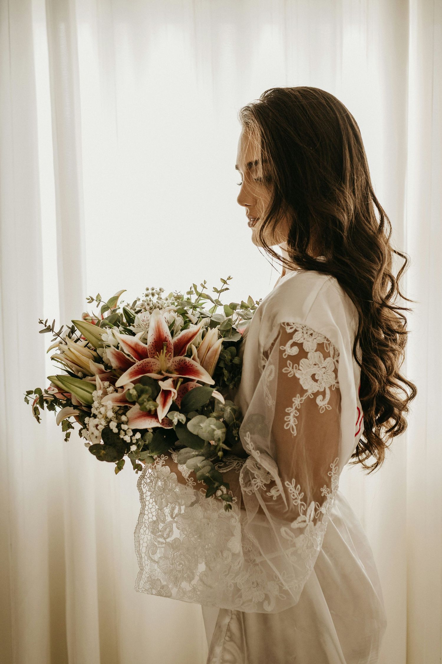 Bride in lace bridal robe holding a stunning bouquet of pink stargazer lilies and greenery.