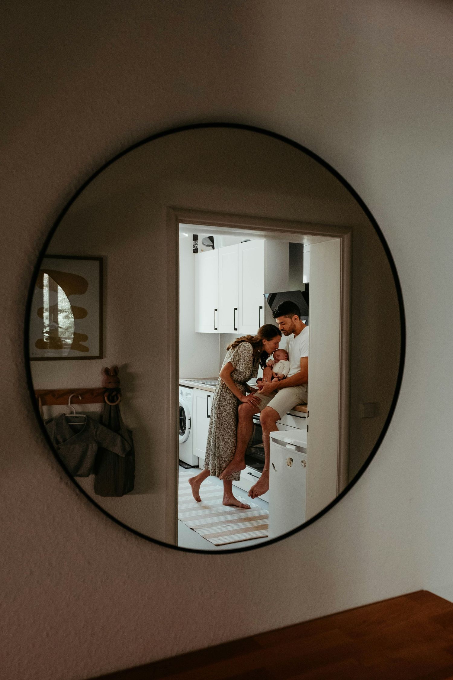 Round mirror reflection of couple with newborn baby in bright modern kitchen with white cabinets.