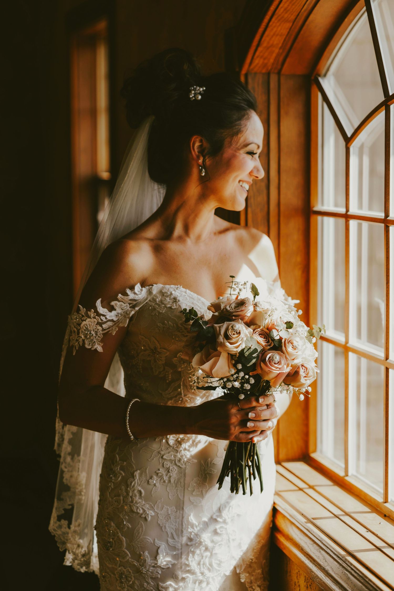 Bride in lace off-shoulder gown holding blush rose bouquet, smiling by sunlit window with veil and hair accessories.