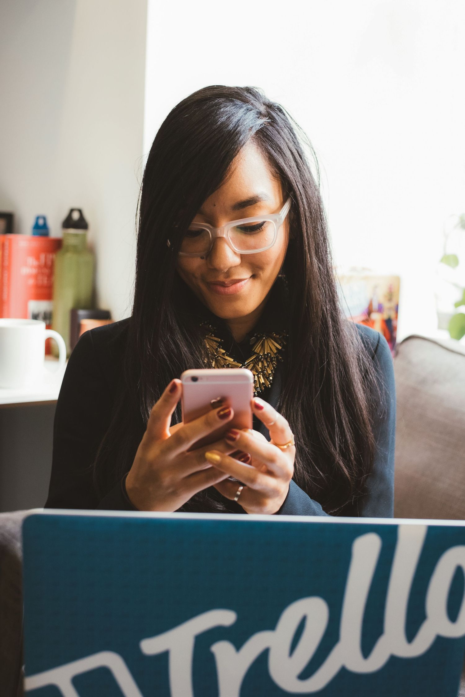 Asian woman with glasses smiling at pink smartphone near Trello laptop in cozy office setting.