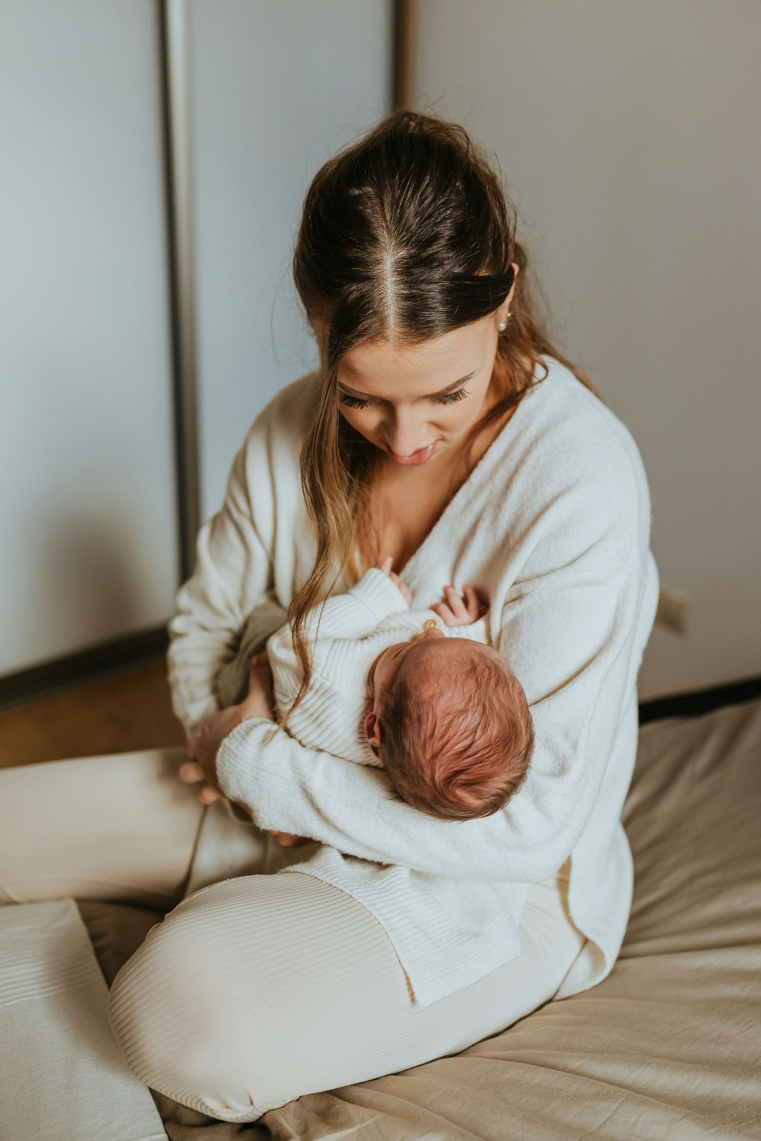 Young mother in white outfit sitting on bed lovingly holding her sleeping newborn baby close.