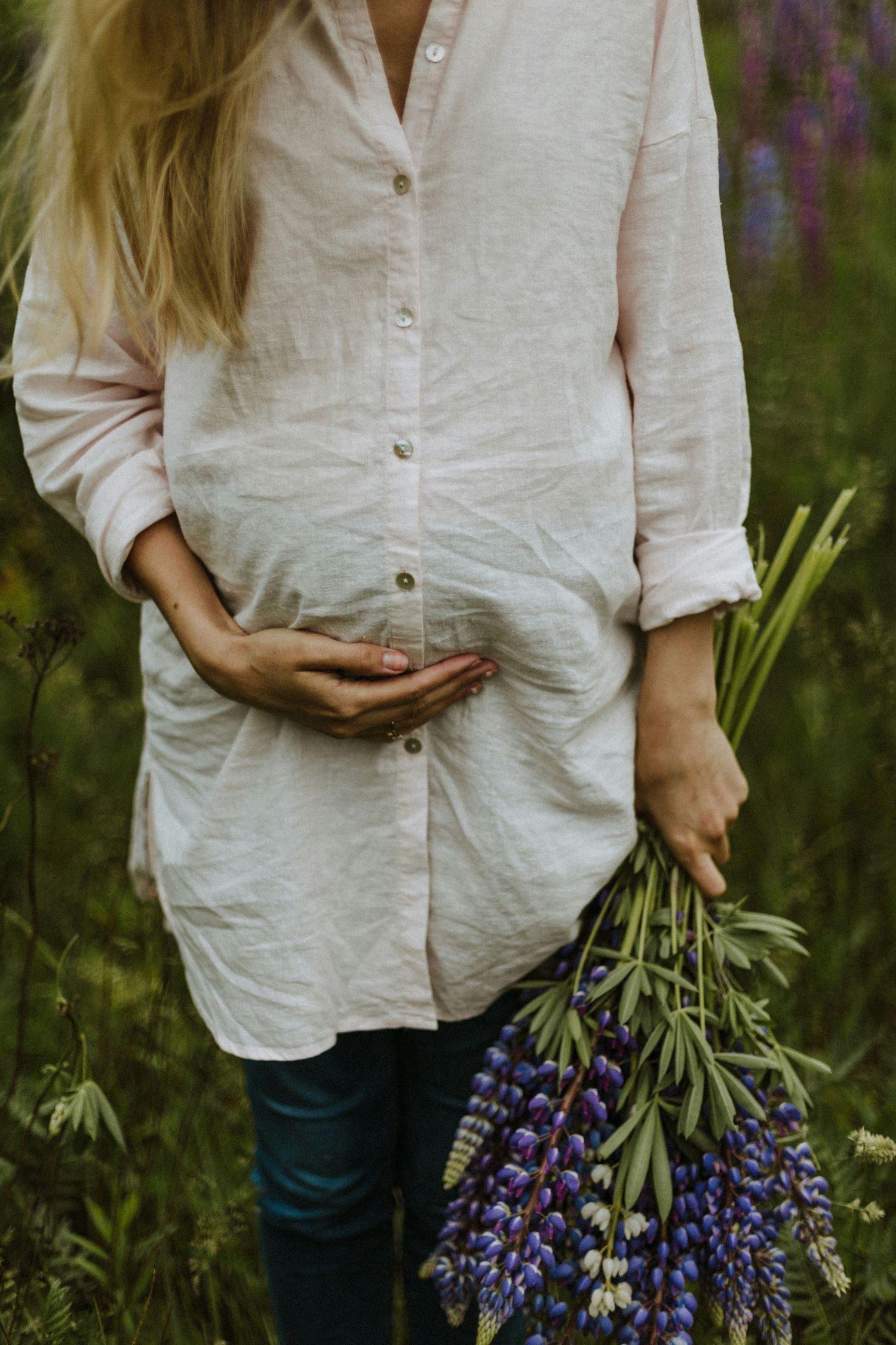 Pregnant woman in linen shirt holding purple lupine flowers in green field.