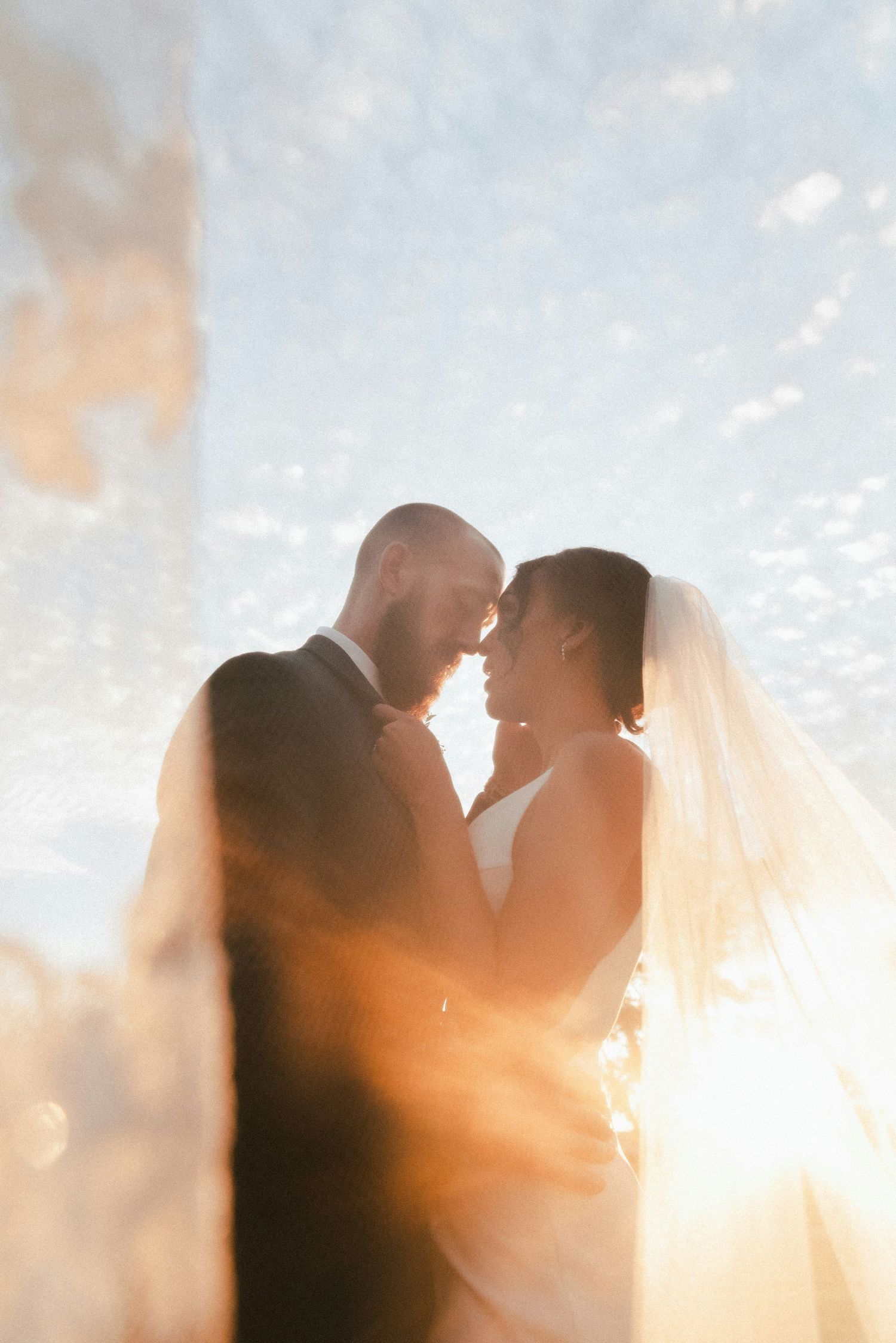 Bride and groom nearly kissing wrapped in veil with stunning golden sunlight and blue sky behind them.