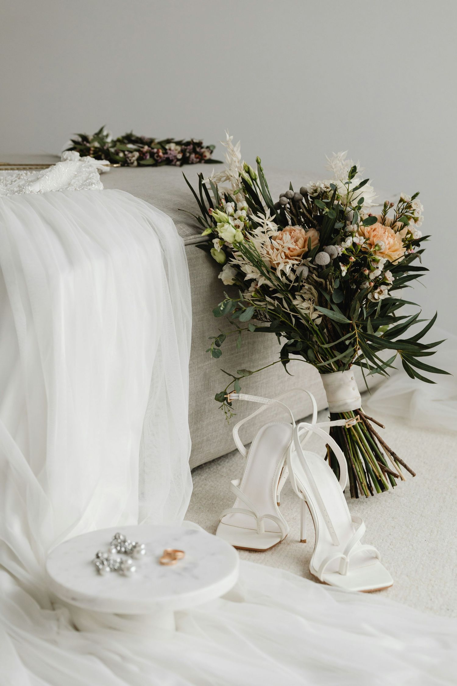 Bridal flat lay with white heeled sandals, a lush bouquet, floral crown, rings, and earrings on white fabric.