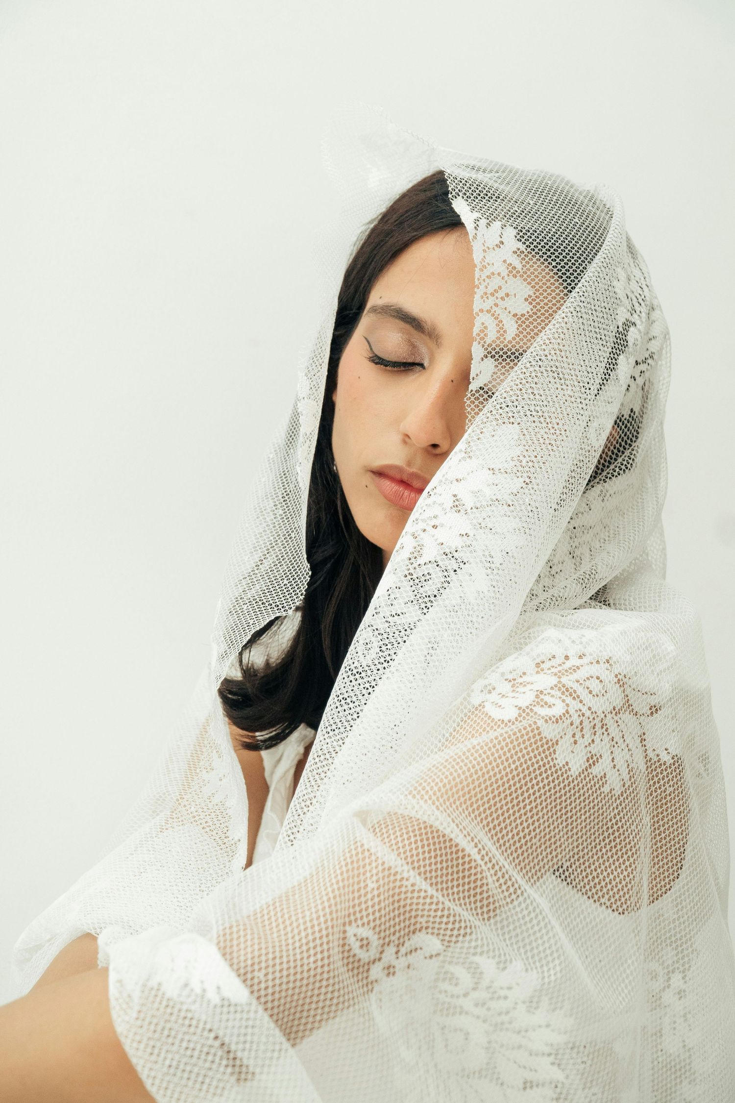 Elegant bride draped in white lace veil with eyes closed on white background.