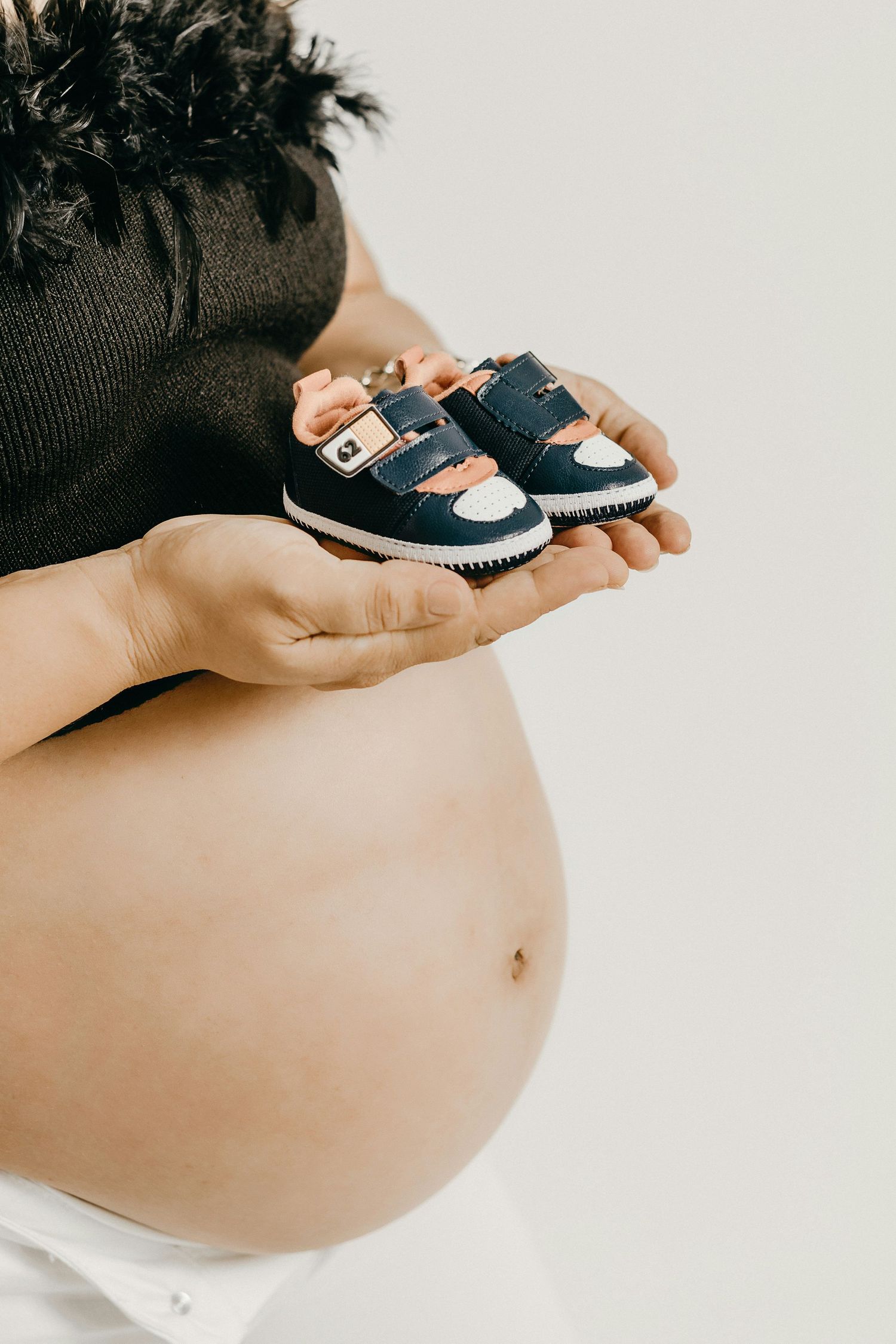 Pregnant woman in black top holding tiny navy blue baby sneakers over her bare bump.