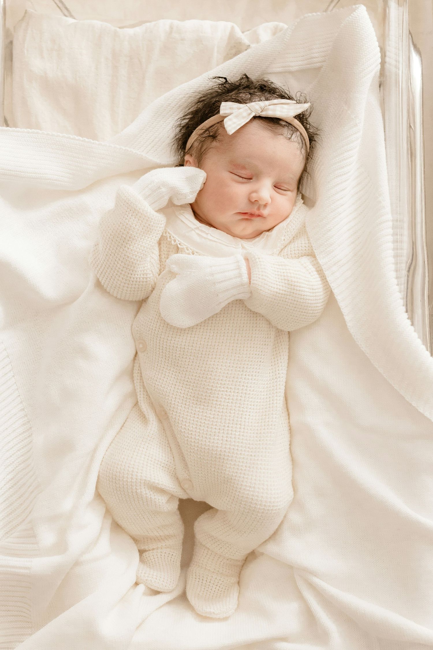 Peaceful newborn baby in cream knit pajamas with a bow headband sleeping in a white crib.