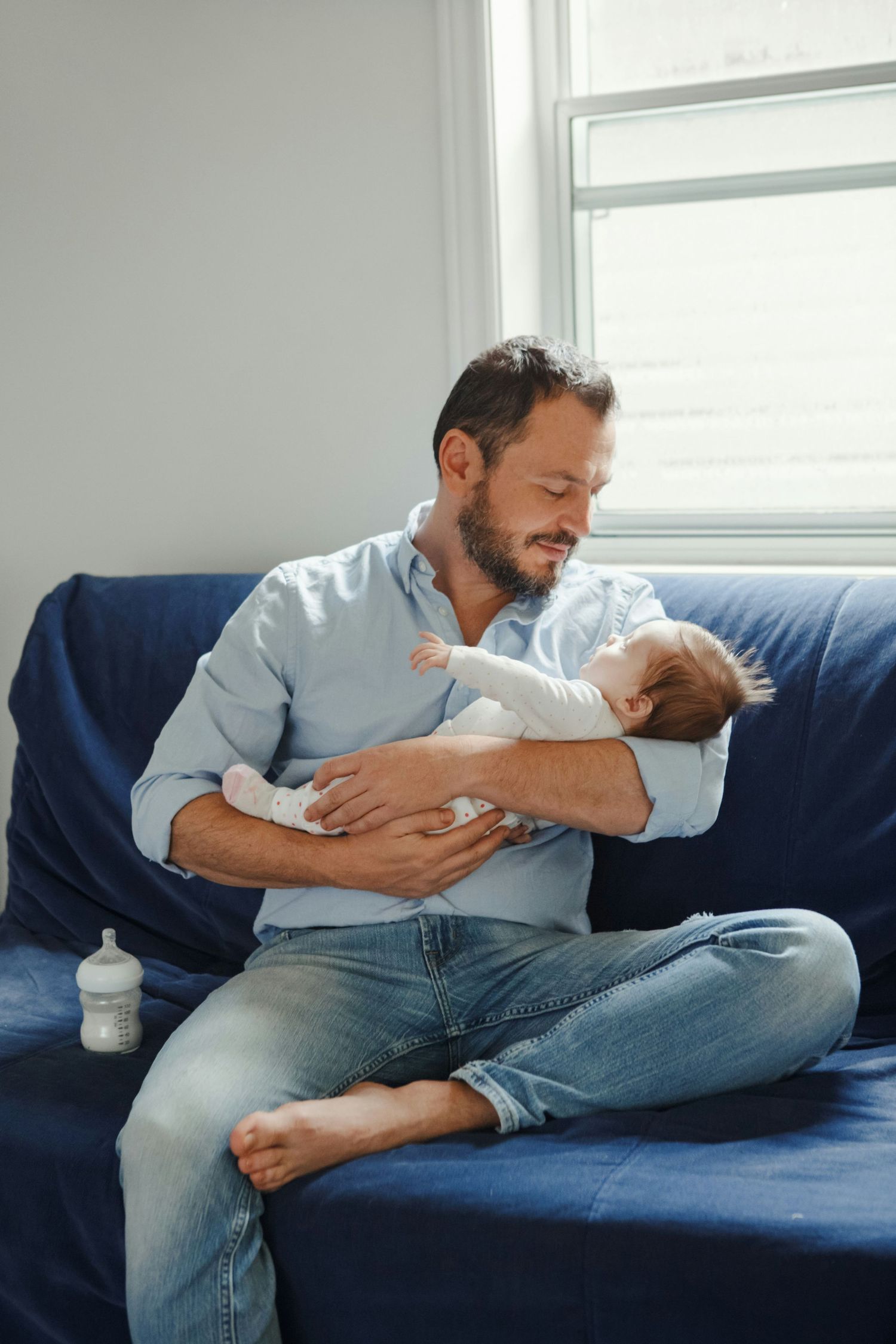 Father in blue shirt sitting cross-legged on navy sofa gently cradling sleeping newborn baby.