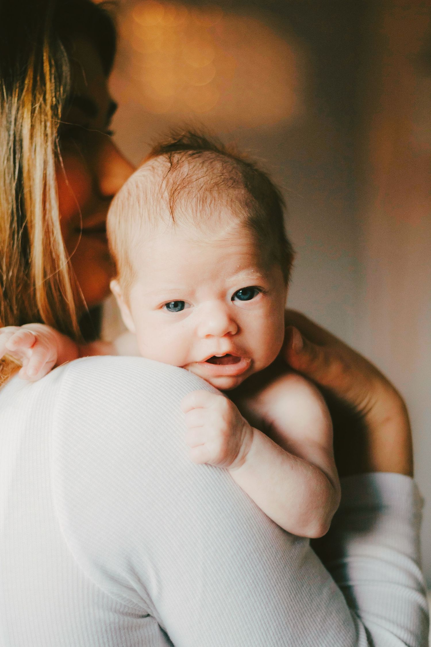 A newborn baby with blue eyes rests on mother's shoulder, looking alert and curious in a warm, softly lit setting.