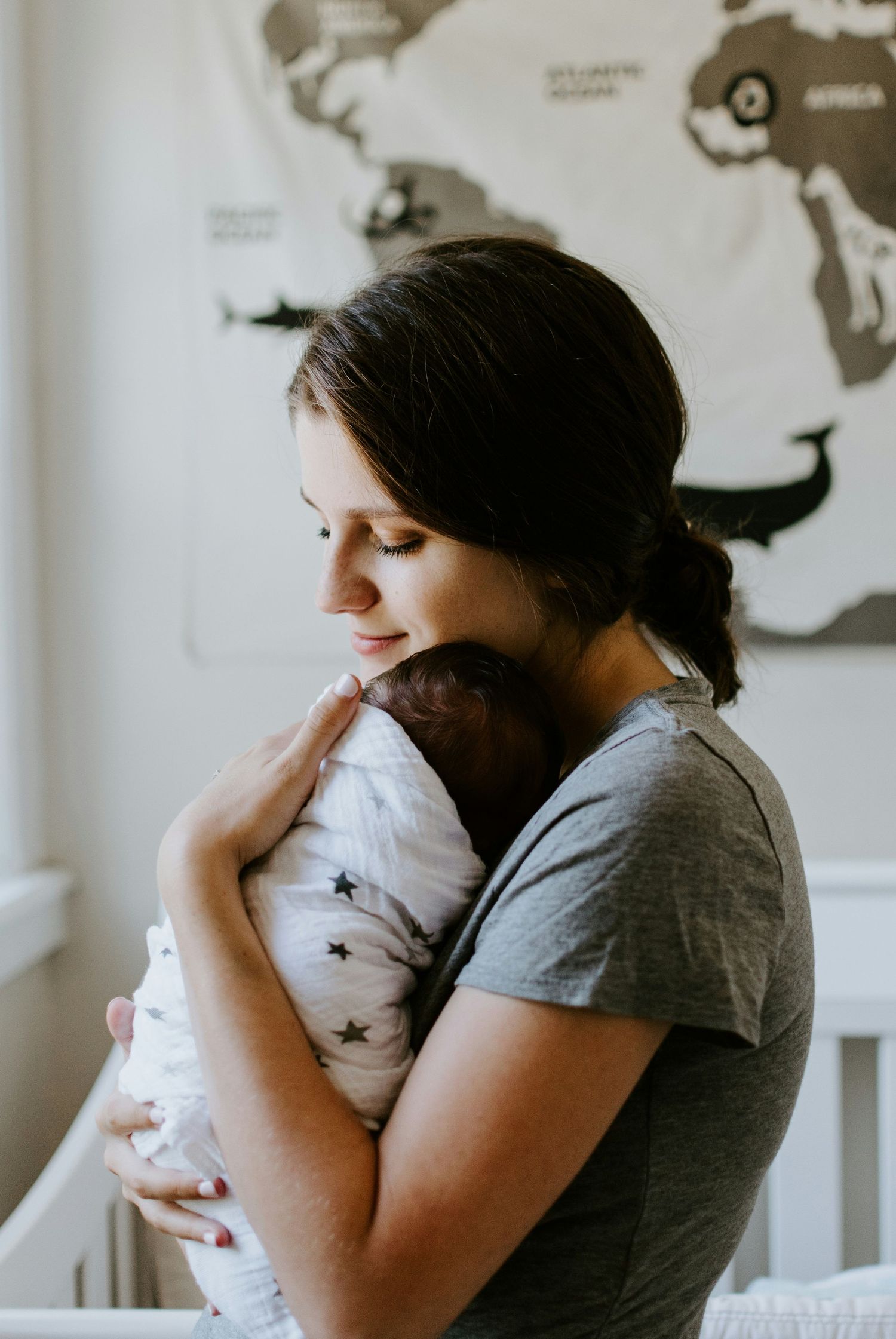 A brunette mother in a gray shirt lovingly cradles her swaddled newborn near a nursery crib.