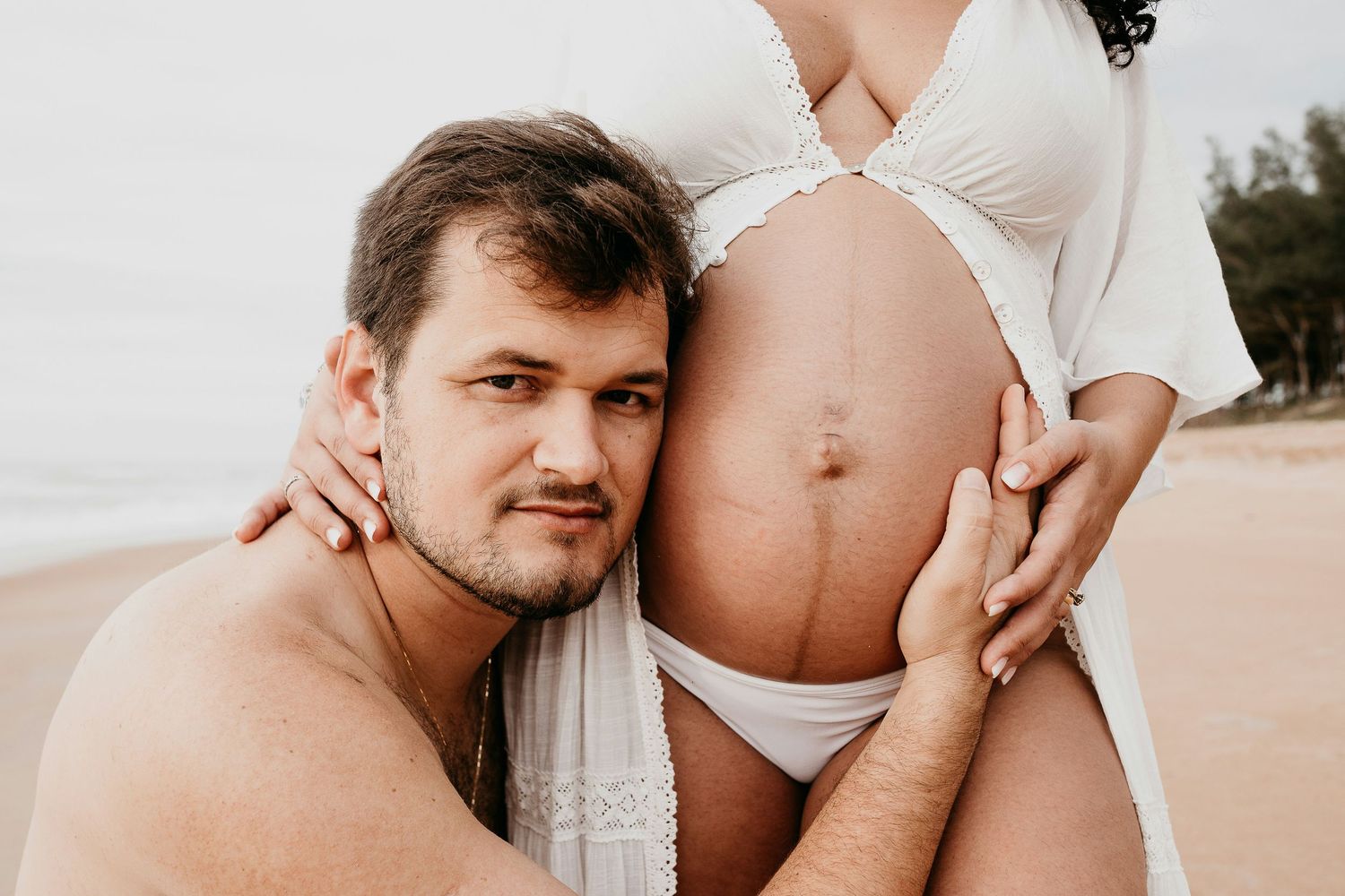 Man resting his head against pregnant woman's bare belly on a sandy beach outdoors.