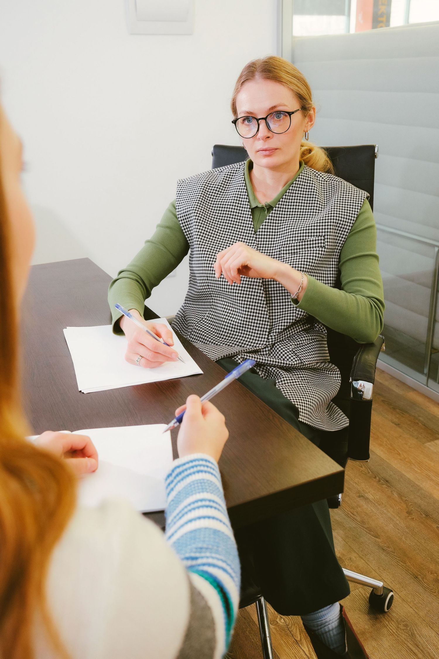 Professional woman in glasses listens attentively during a business meeting, taking notes at an office desk.