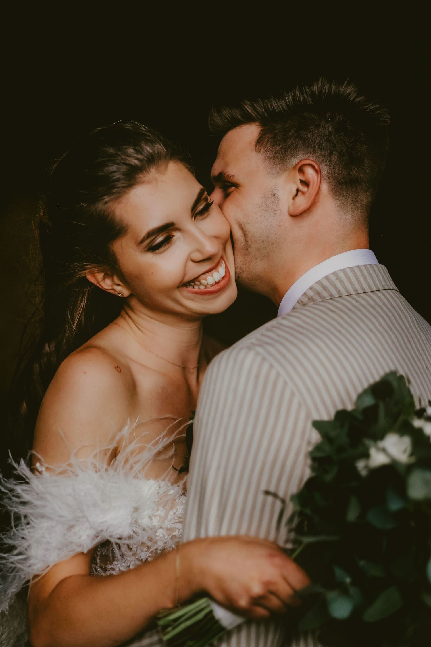 A smiling bride in a feathered white dress receives a kiss on the cheek from a groom in a striped suit holding a bouquet.