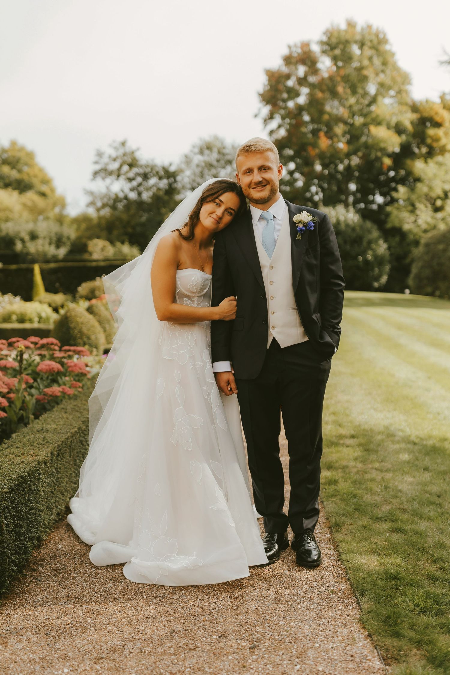A bride in a white gown and veil leans on a groom in a dark suit, posing in a formal garden.