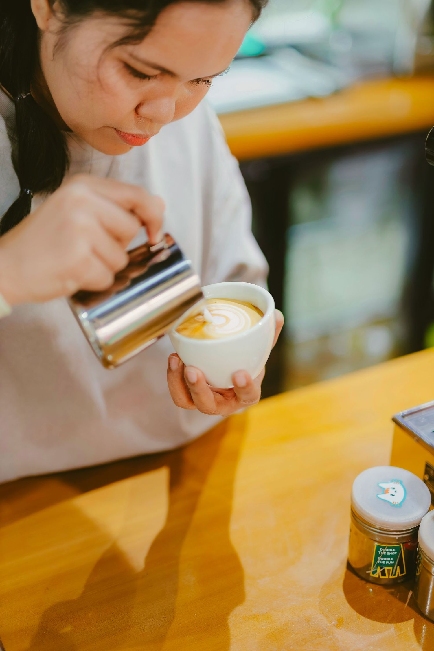 Barista pouring steamed milk into a latte art coffee cup on a wooden counter in a cafe.