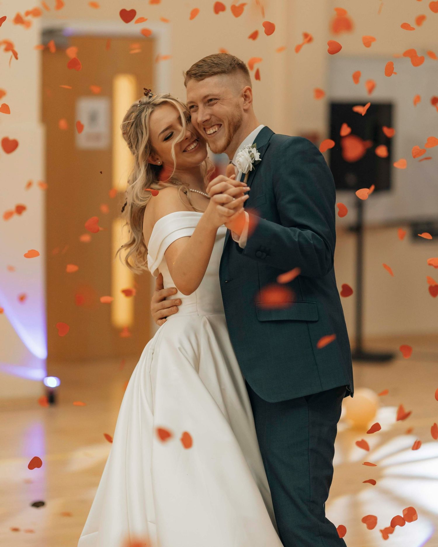 Happy newlyweds dancing amid falling red heart confetti, groom in teal suit, bride in white gown.