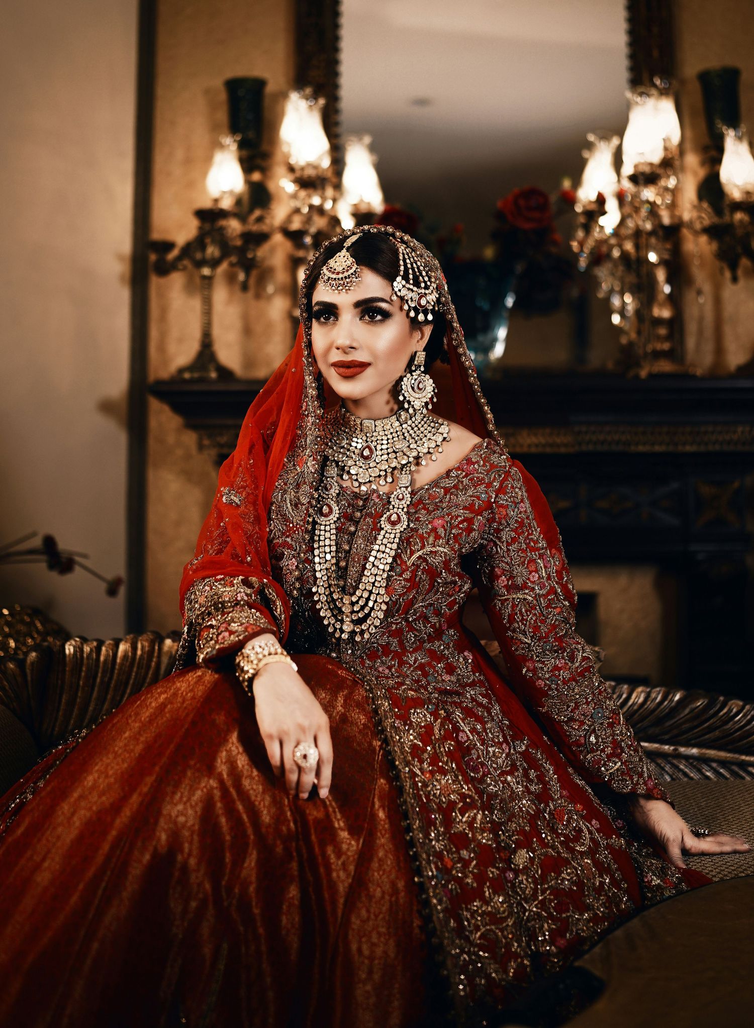Pakistani bride in ornate red bridal lehenga with gold embroidery and traditional jewelry indoors.