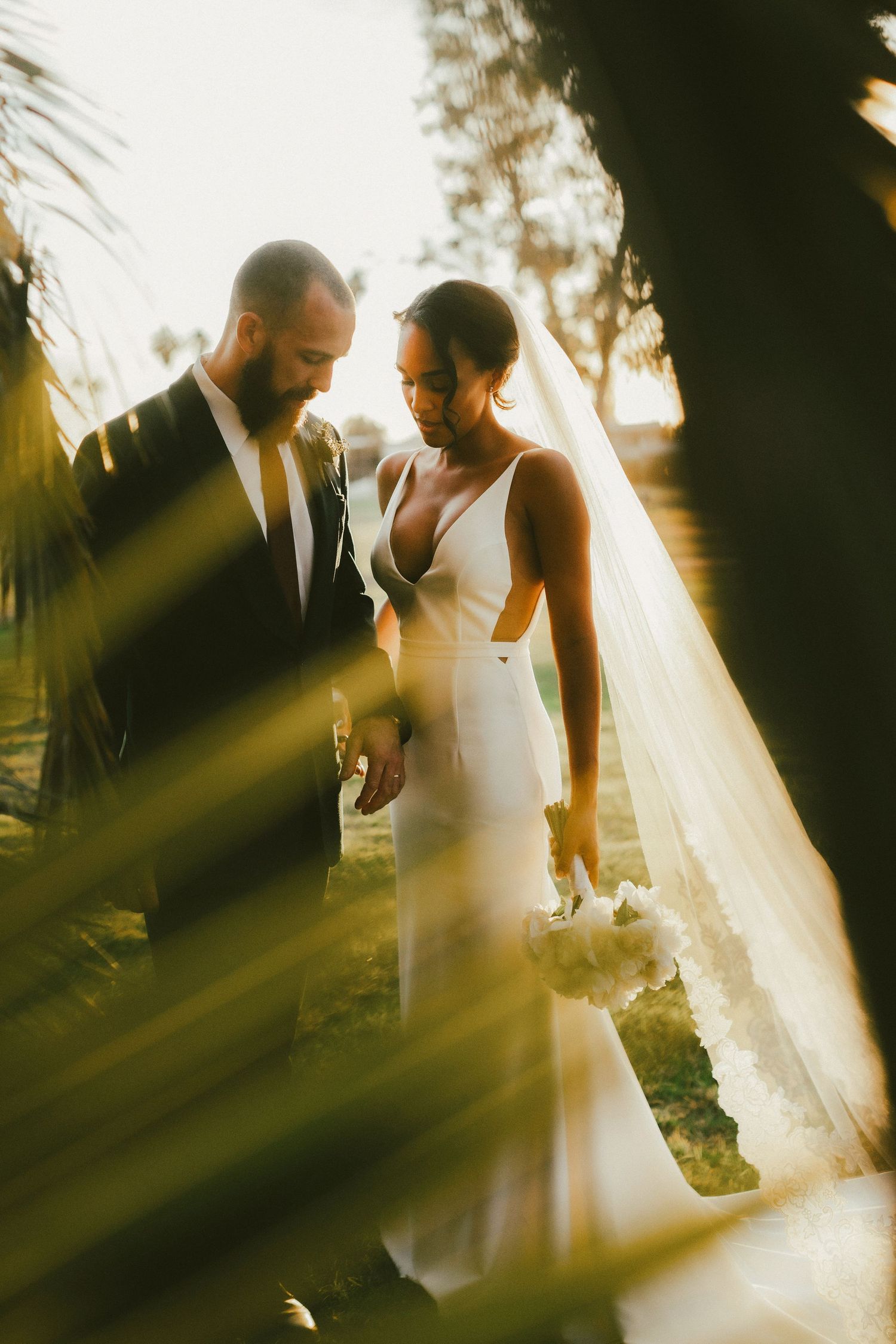 A bride and groom share a romantic moment in golden sunlight, framed by lush greenery during their outdoor wedding.