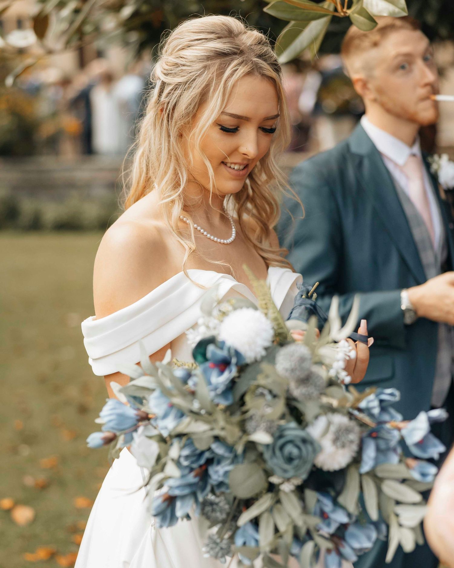 Bride in off-shoulder white gown holding blue floral bouquet, smiling outdoors at wedding ceremony.