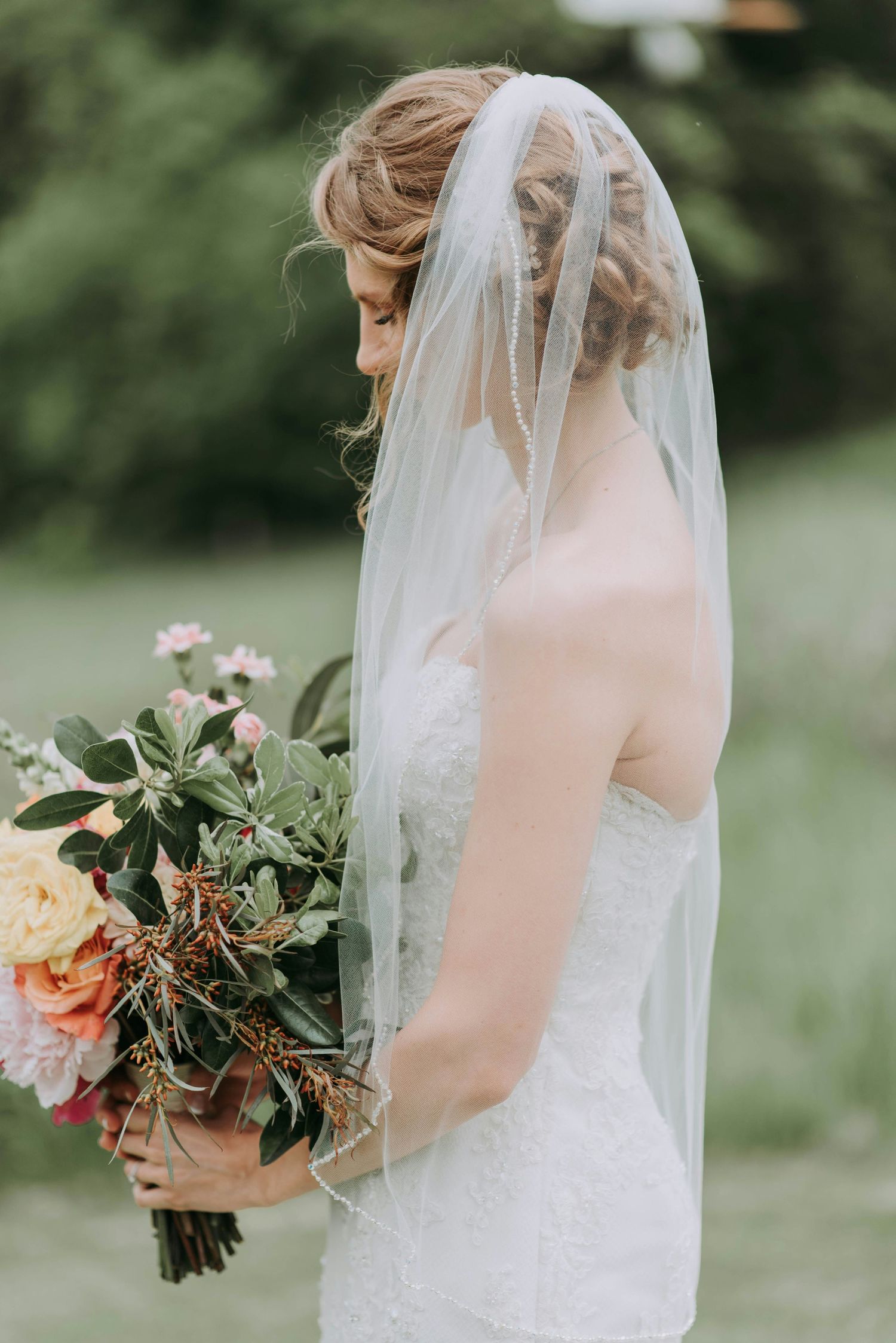 Bride in white strapless lace gown with pearl-edged veil holding colorful floral bouquet.