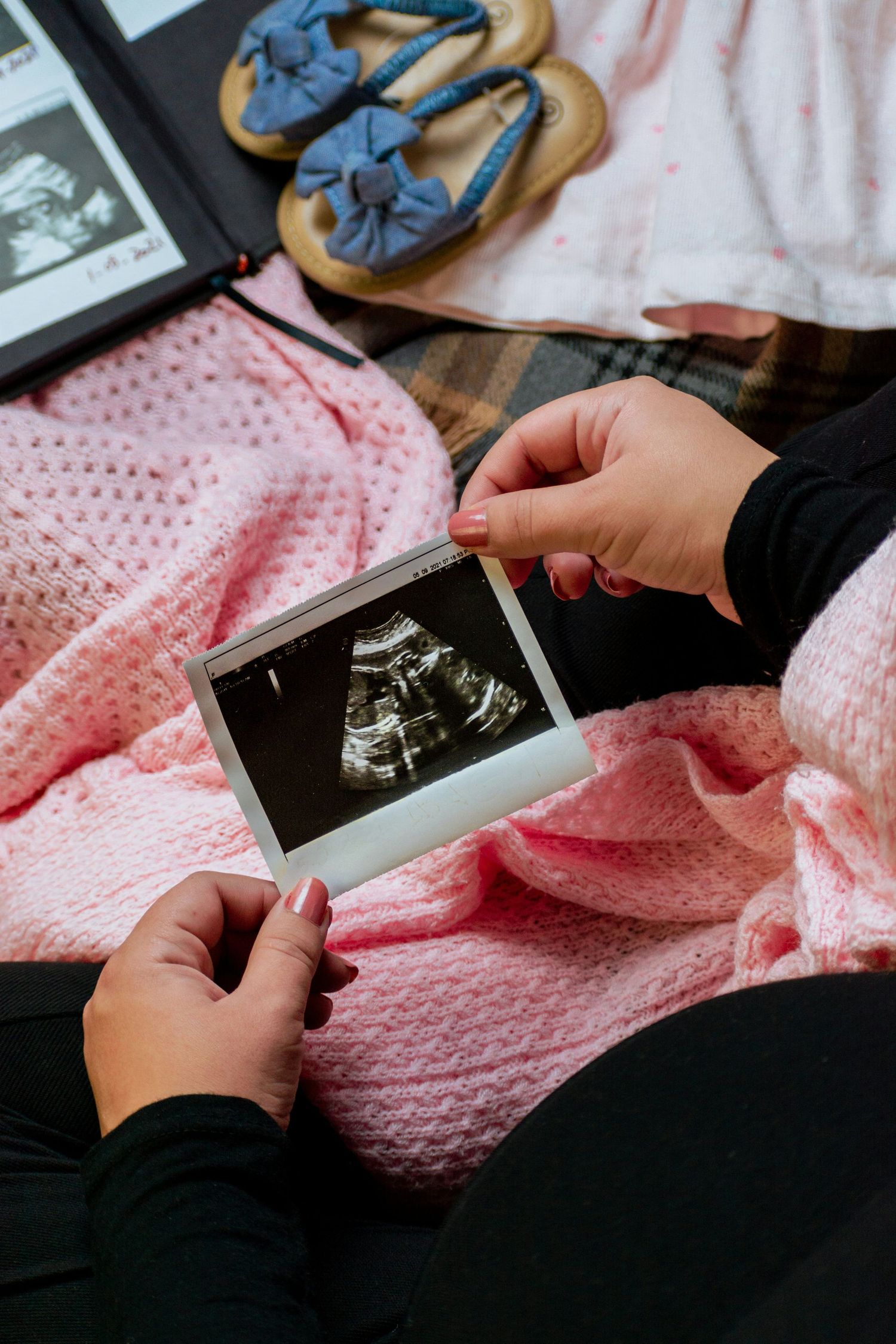 Pregnant woman holding ultrasound photo on pink blanket with baby shoes nearby.