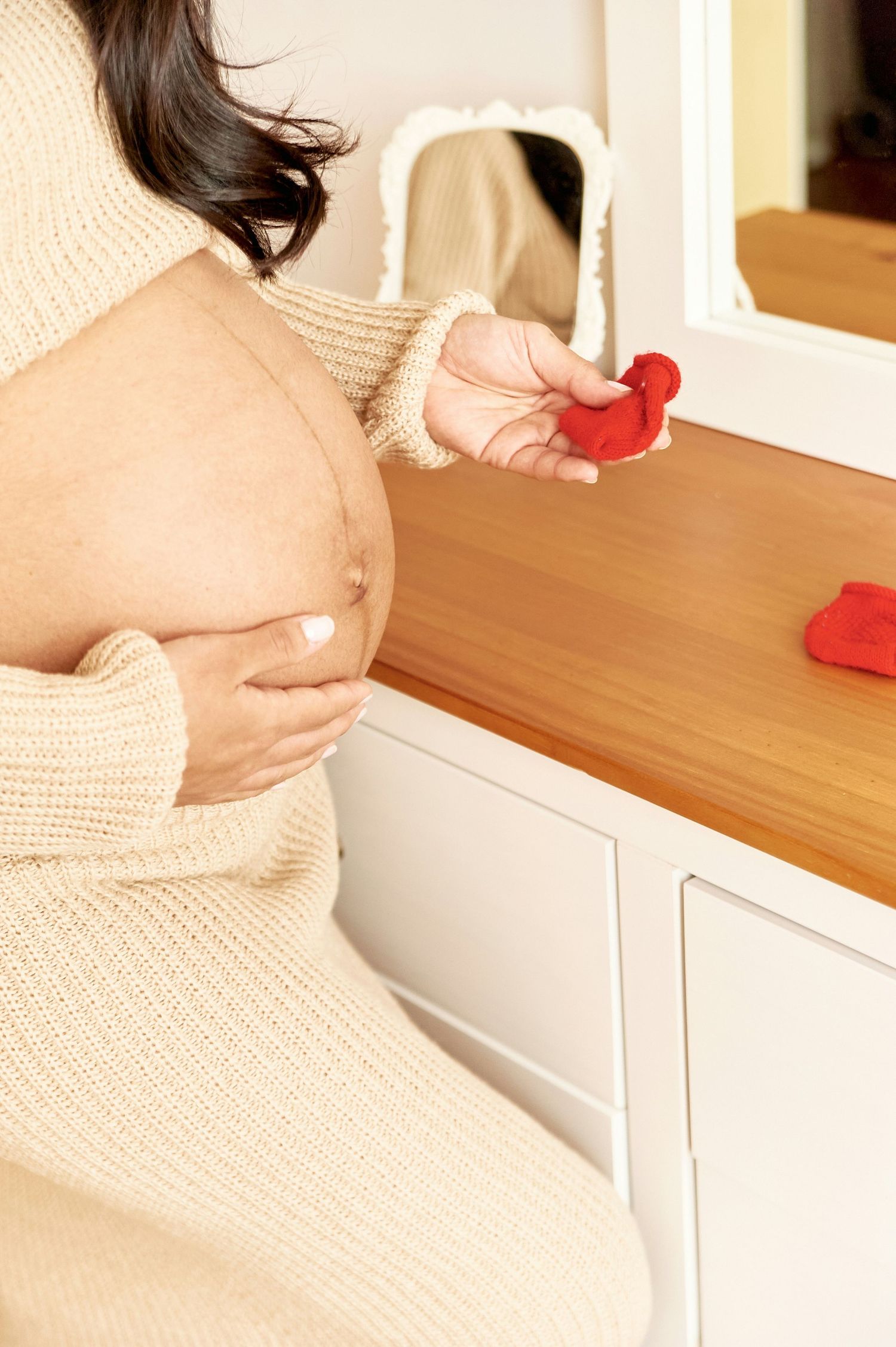 Pregnant woman in beige sweater holding a small red clay figurine at a wooden dresser.