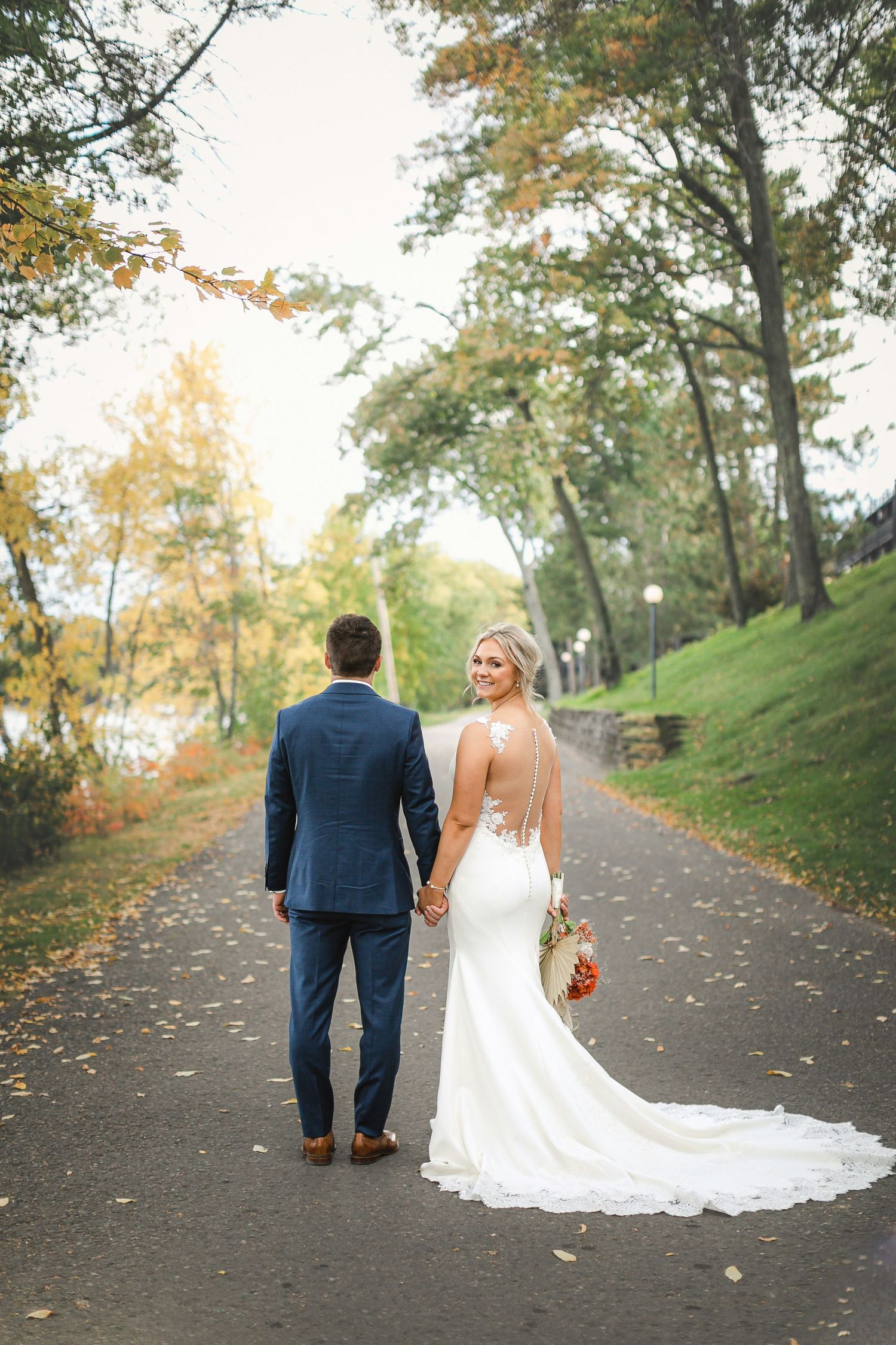 Bride and groom walk hand in hand down a tree-lined path in autumn, bride's gown trailing behind them.