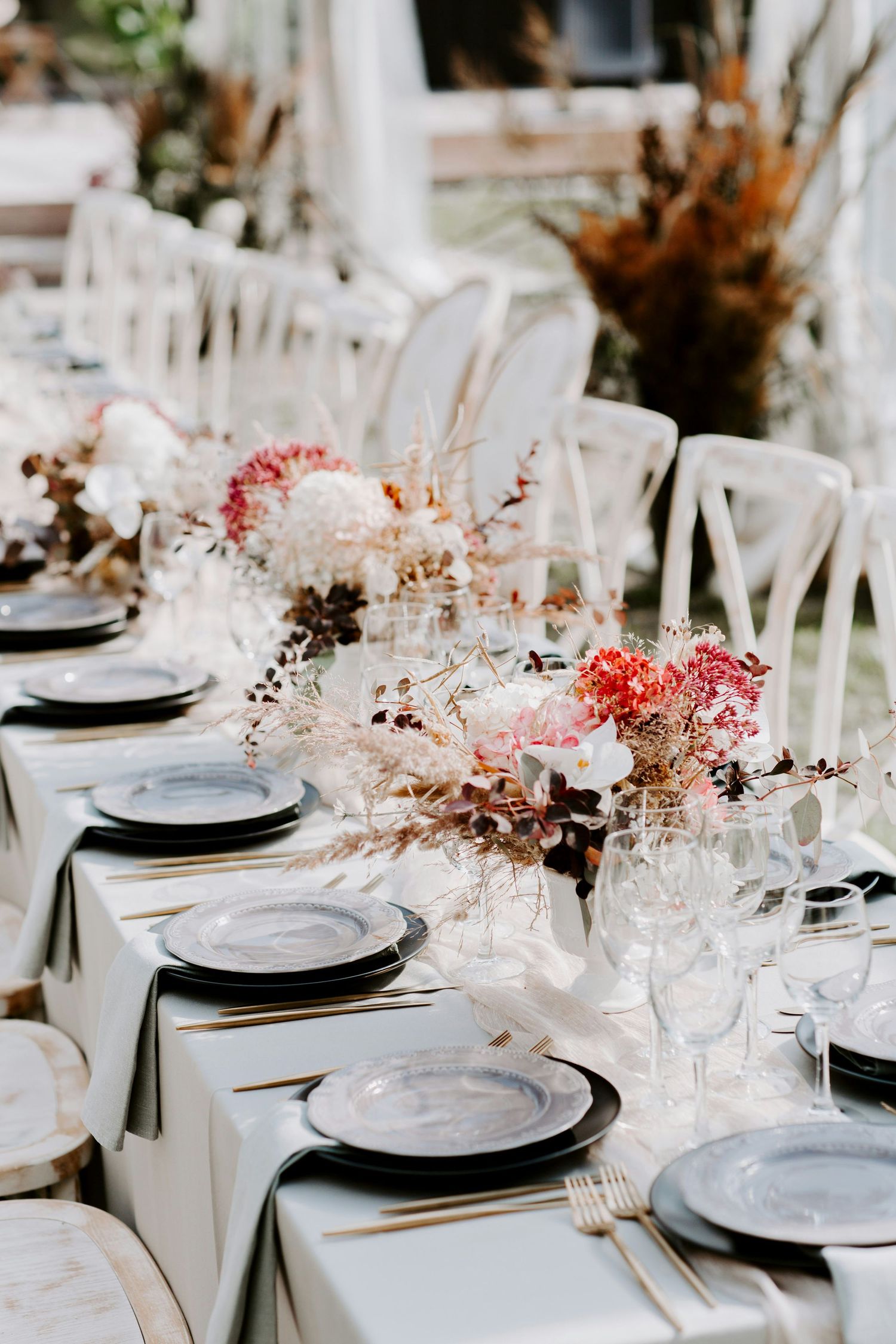 Elegant outdoor wedding reception table with dark plates, gold cutlery, and dried floral centerpieces.