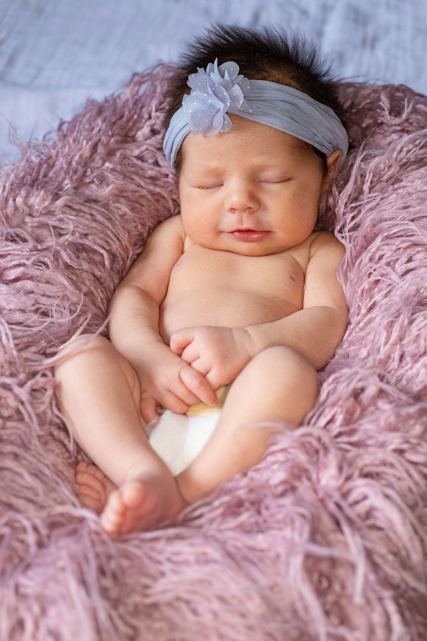 Sleeping newborn baby girl with floral headband resting on pink fluffy fur blanket.