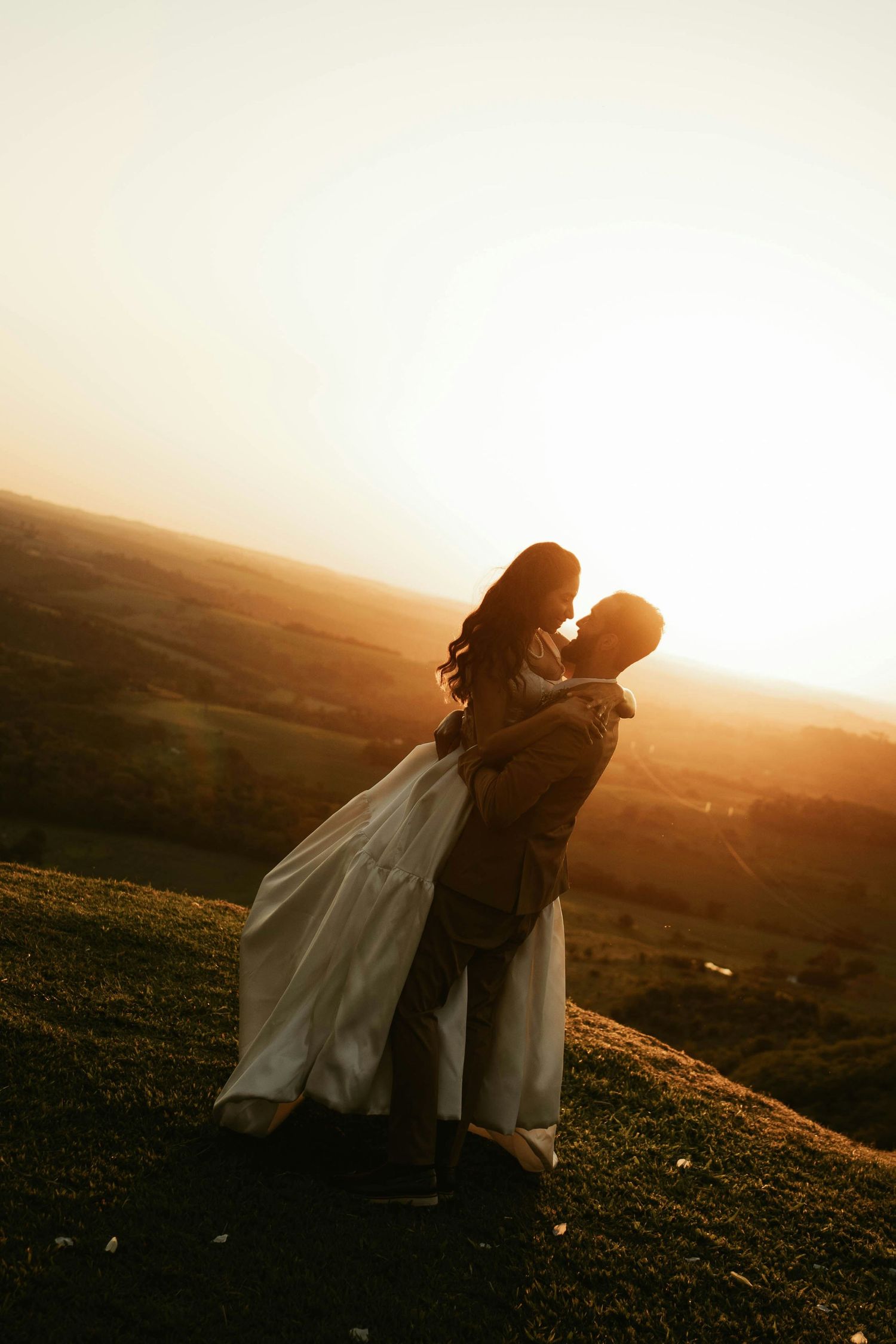 A groom lifts his bride in a flowing white dress at sunset on a scenic hilltop landscape.
