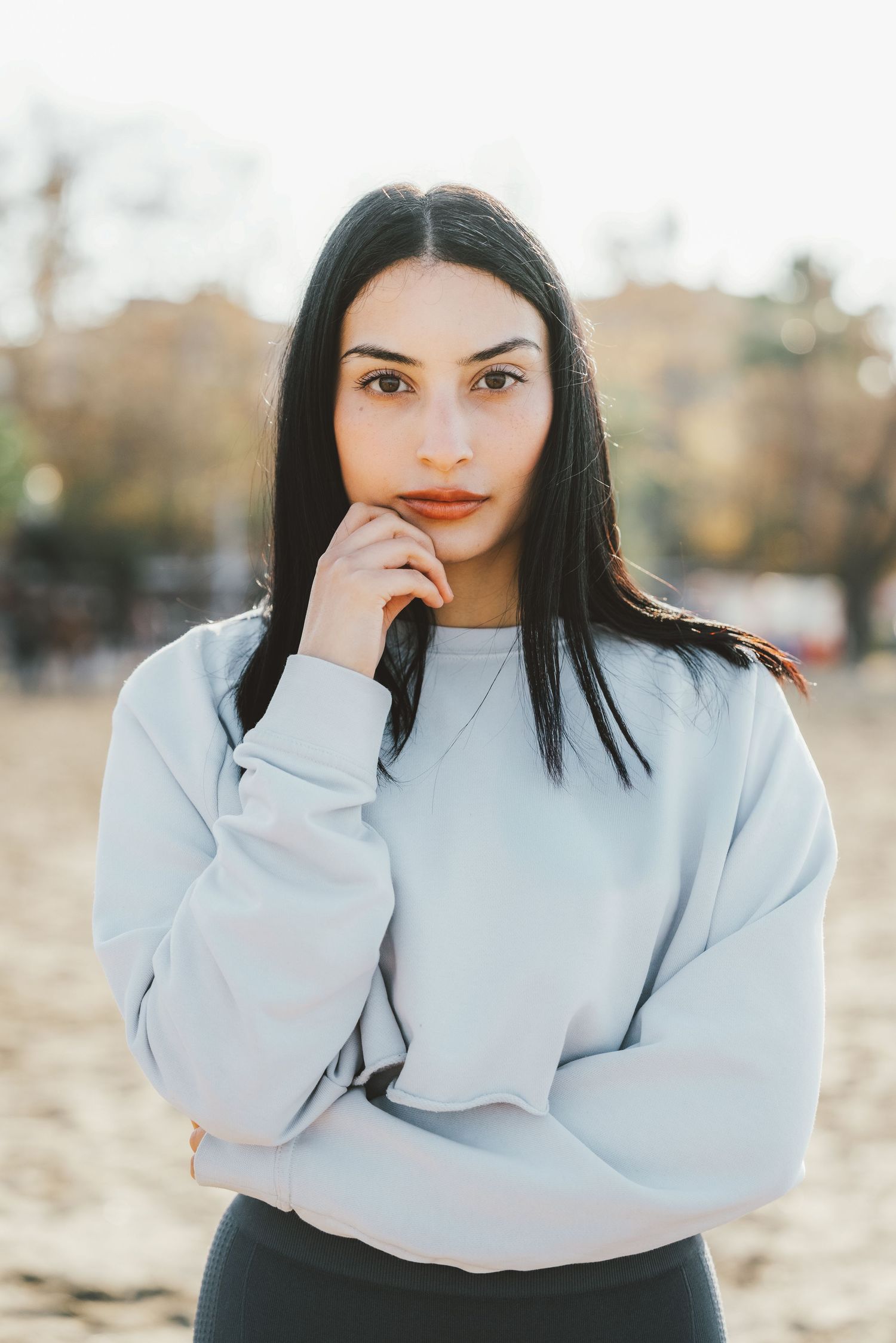 Young woman with straight black hair in white sweatshirt posing thoughtfully outdoors at golden hour.