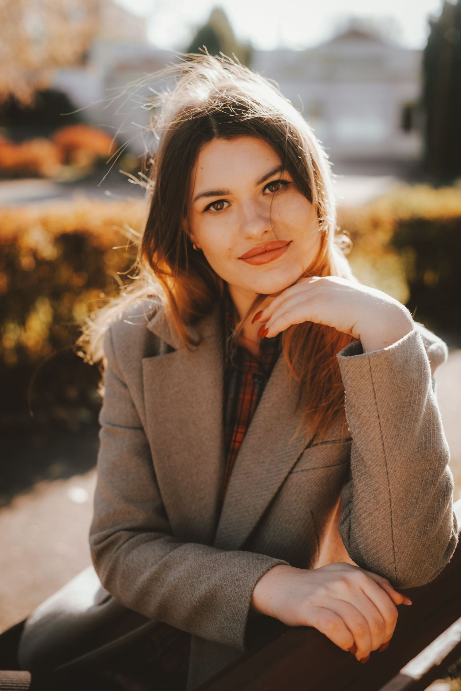 Young brunette woman in gray blazer sitting outdoors in warm sunlight smiling.