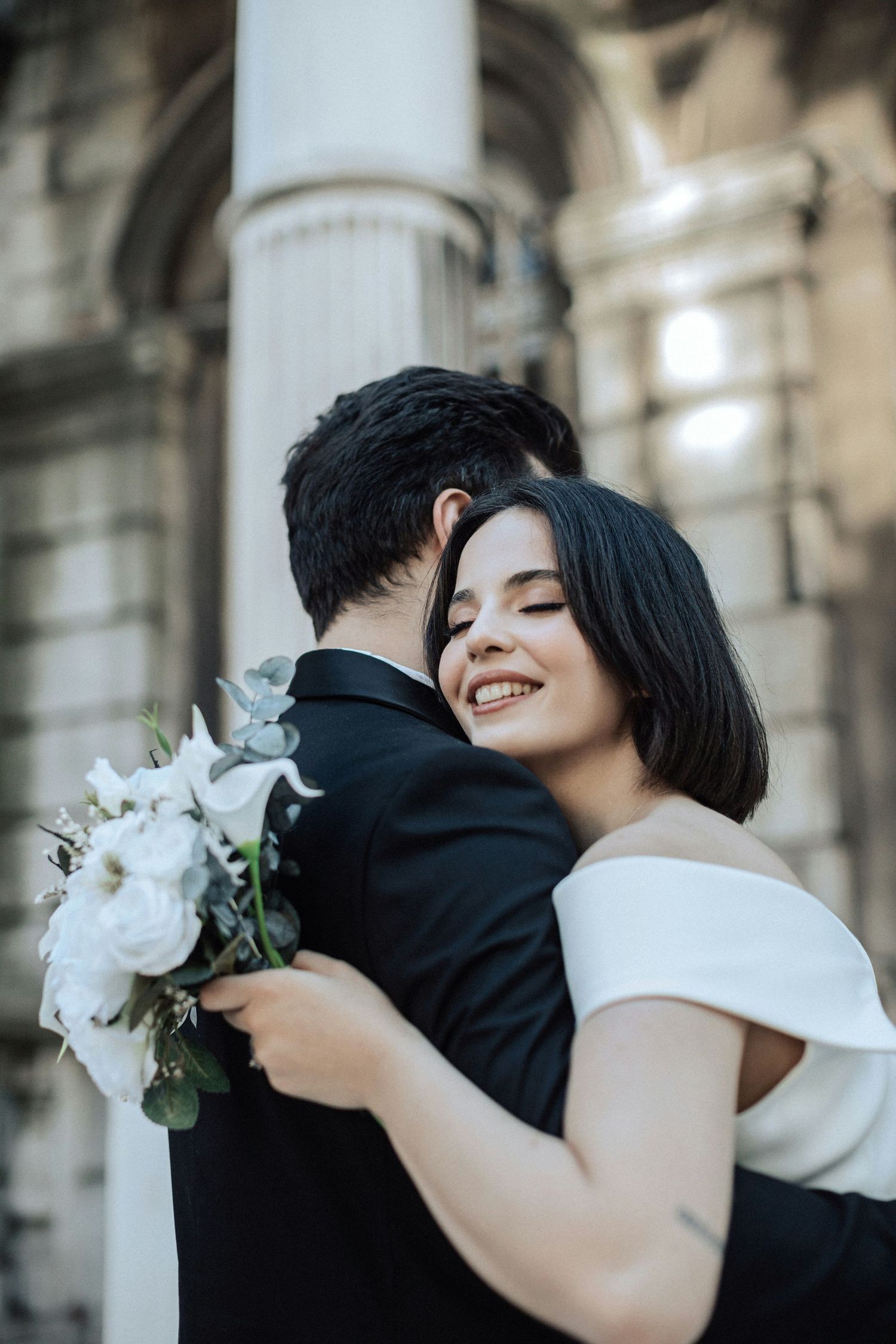 Joyful dark-haired bride in off-shoulder white dress embraces groom while clutching white rose bouquet outdoors.