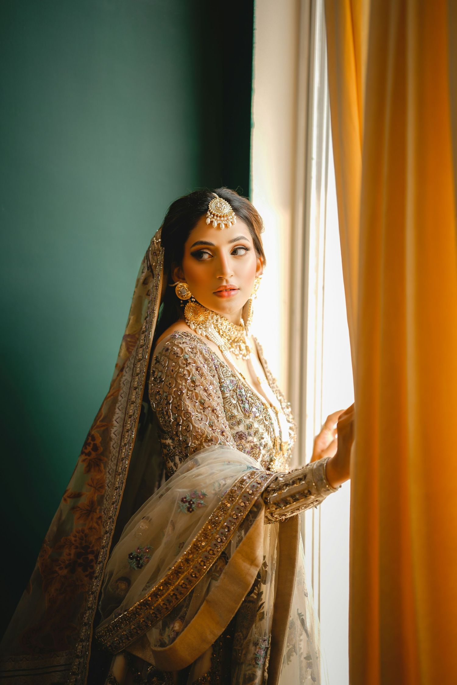 Elegant bride in ivory embroidered Pakistani bridal outfit standing by a sunlit window.