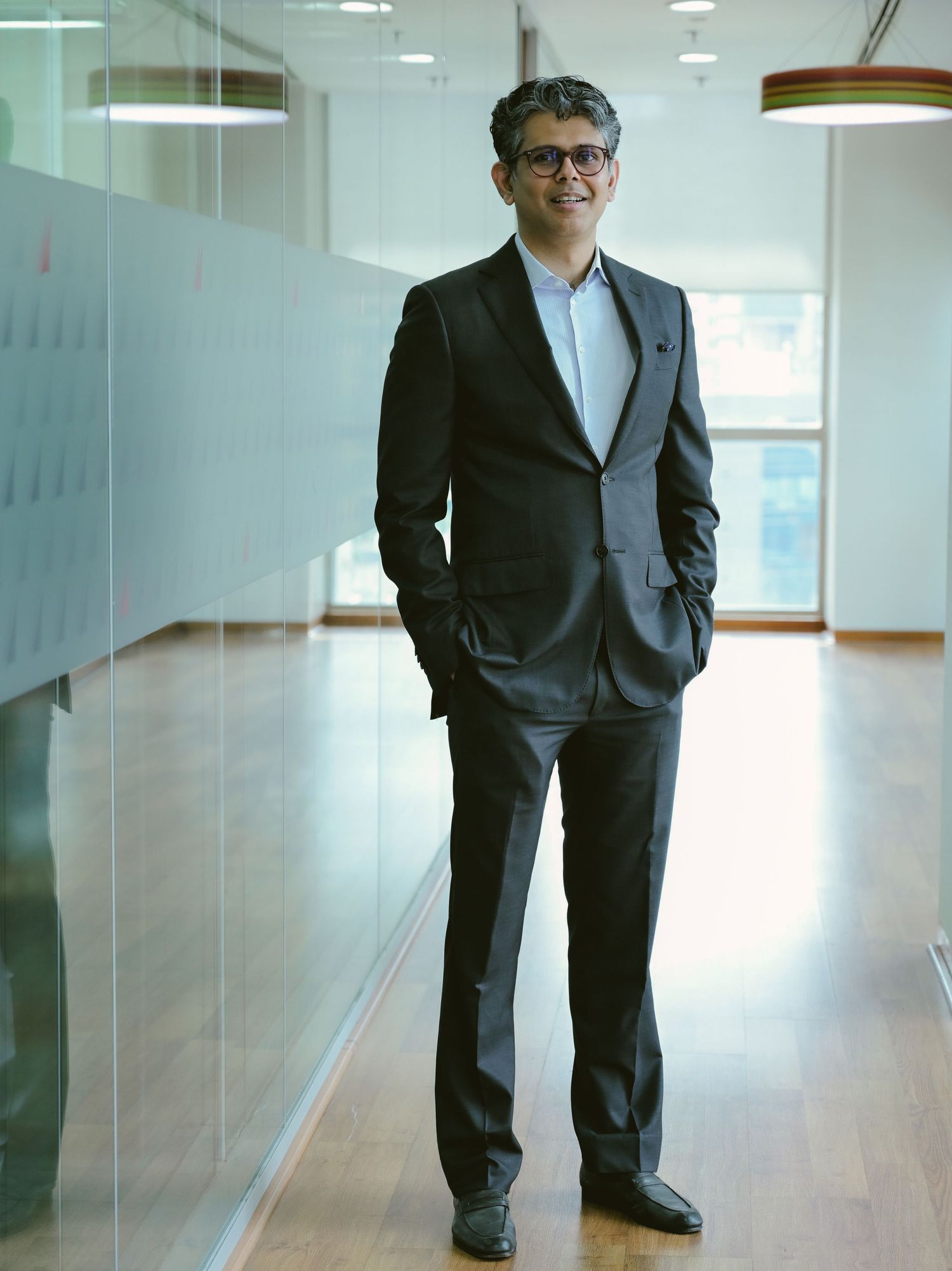 Professional man in dark suit standing confidently in modern office corridor with glass walls and wooden floors.