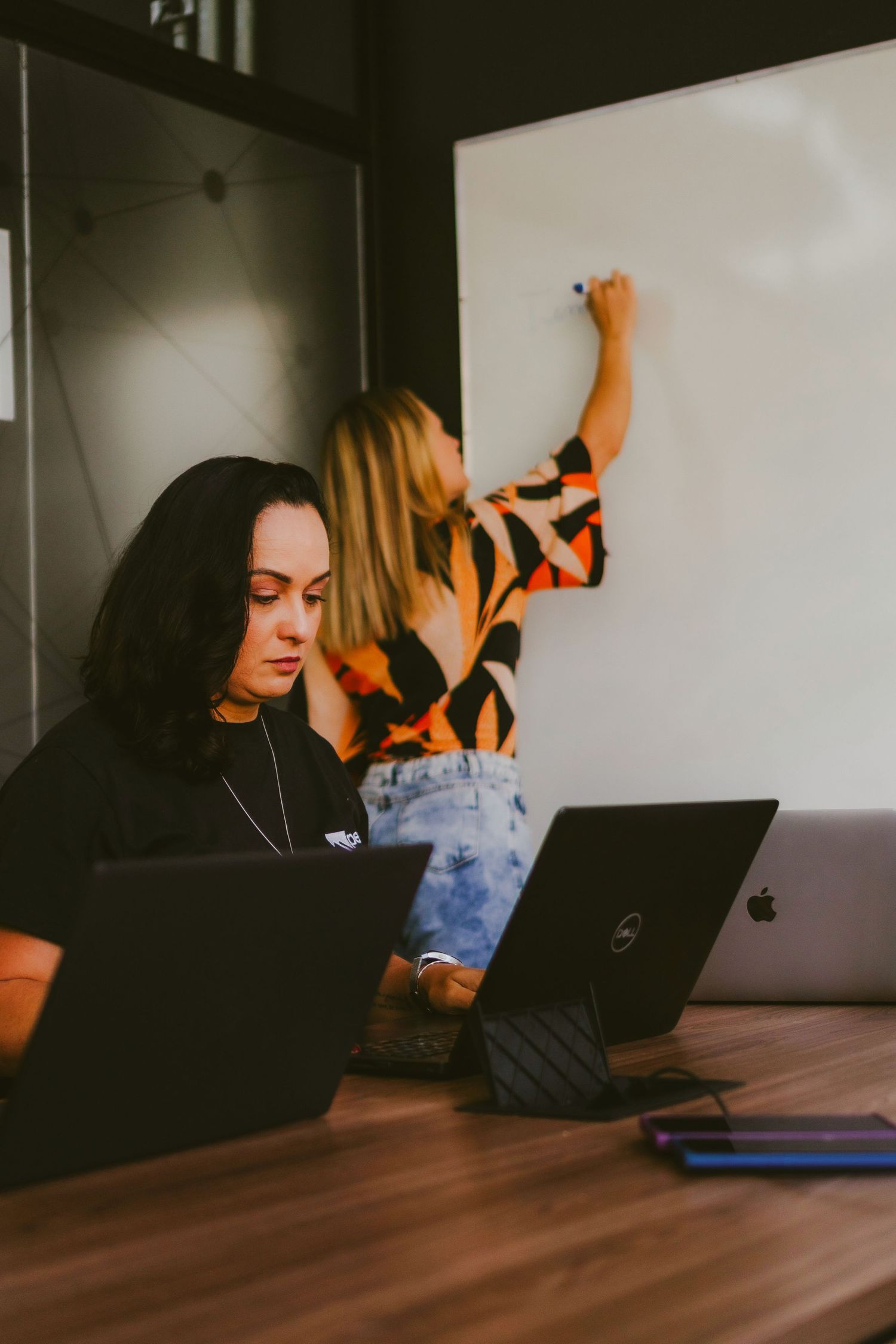 Two women collaborating in office, one writing on whiteboard while other works on laptop and tablet at wooden desk.