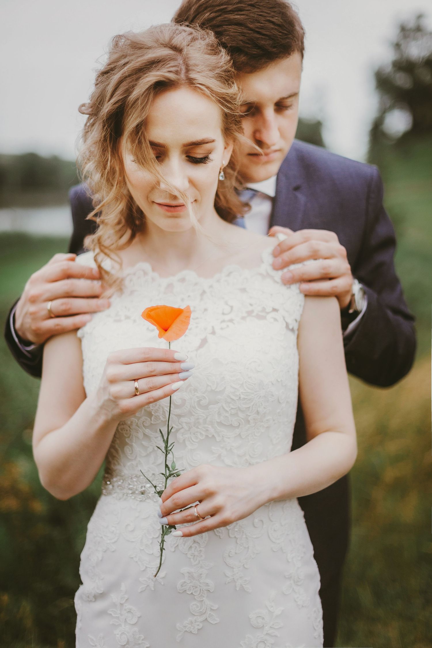 Bride in lace wedding dress holds orange poppy flower while groom in blue suit embraces her from behind outdoors.