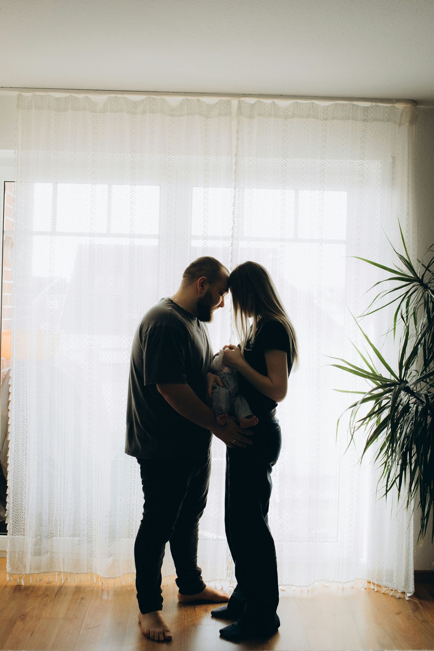 Silhouette of couple standing by bright window together holding newborn baby indoors.
