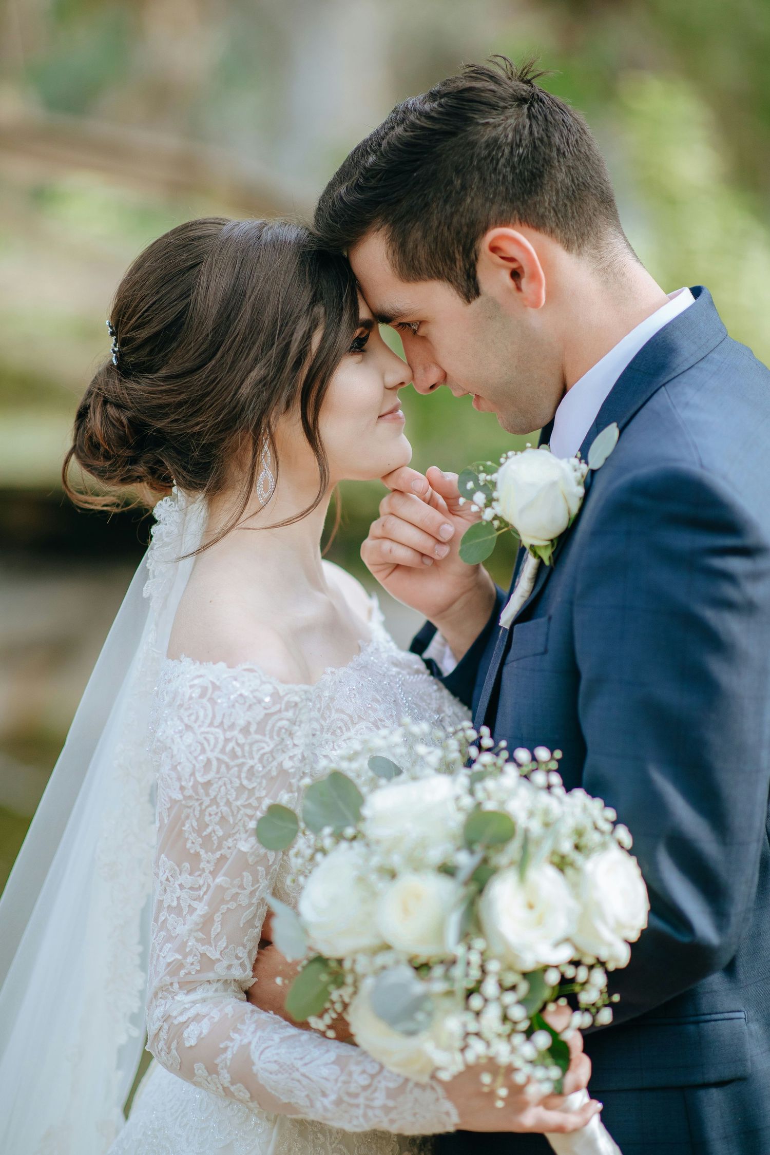 Bride in lace gown and groom in blue suit touching foreheads romantically while holding white rose bouquet.