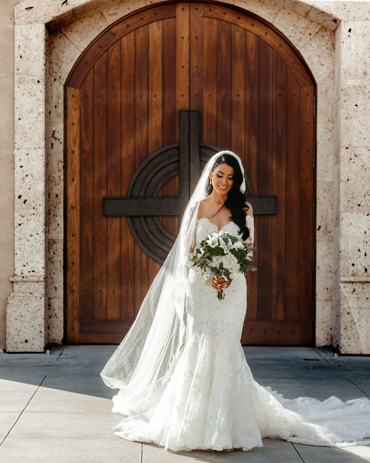 Bride in lace mermaid wedding gown with cathedral veil holding white bouquet before wooden church doors.